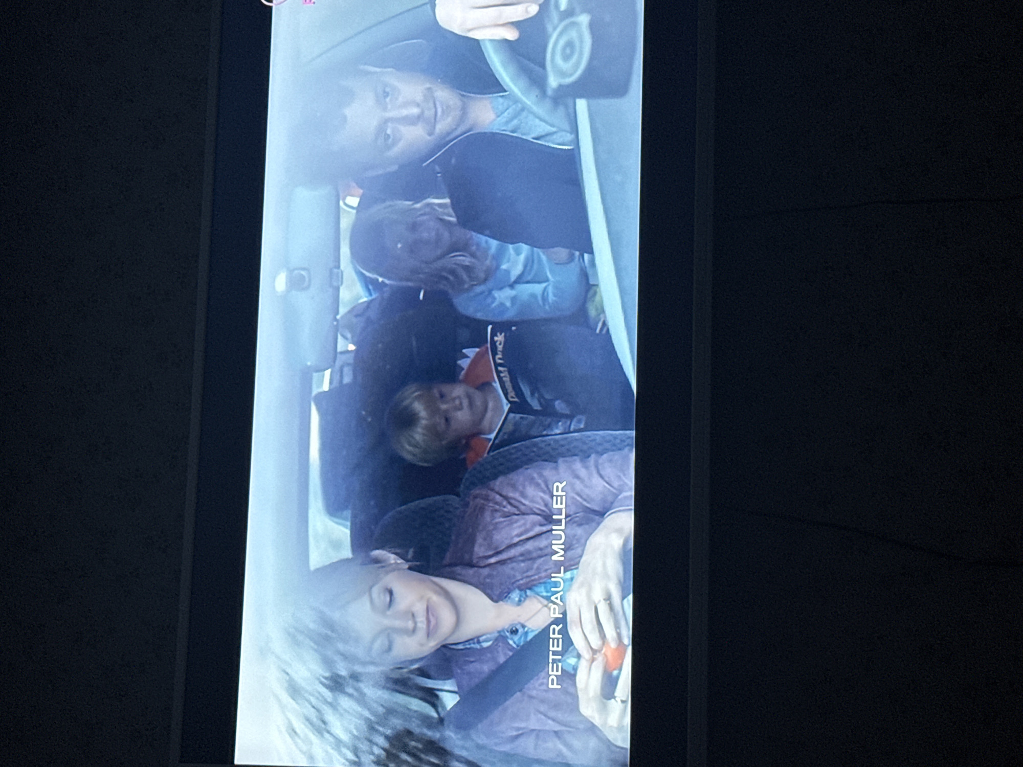The image shows a family in a car, with a man driving, a woman in the passenger seat, and three children in the back. The woman is wearing a seatbelt and holding a small orange object in her hands. The driver is looking at the camera. The context suggests the family is on a trip, possibly a road trip. There is no evidence to suggest this image was taken in Mytishchi, Russia.