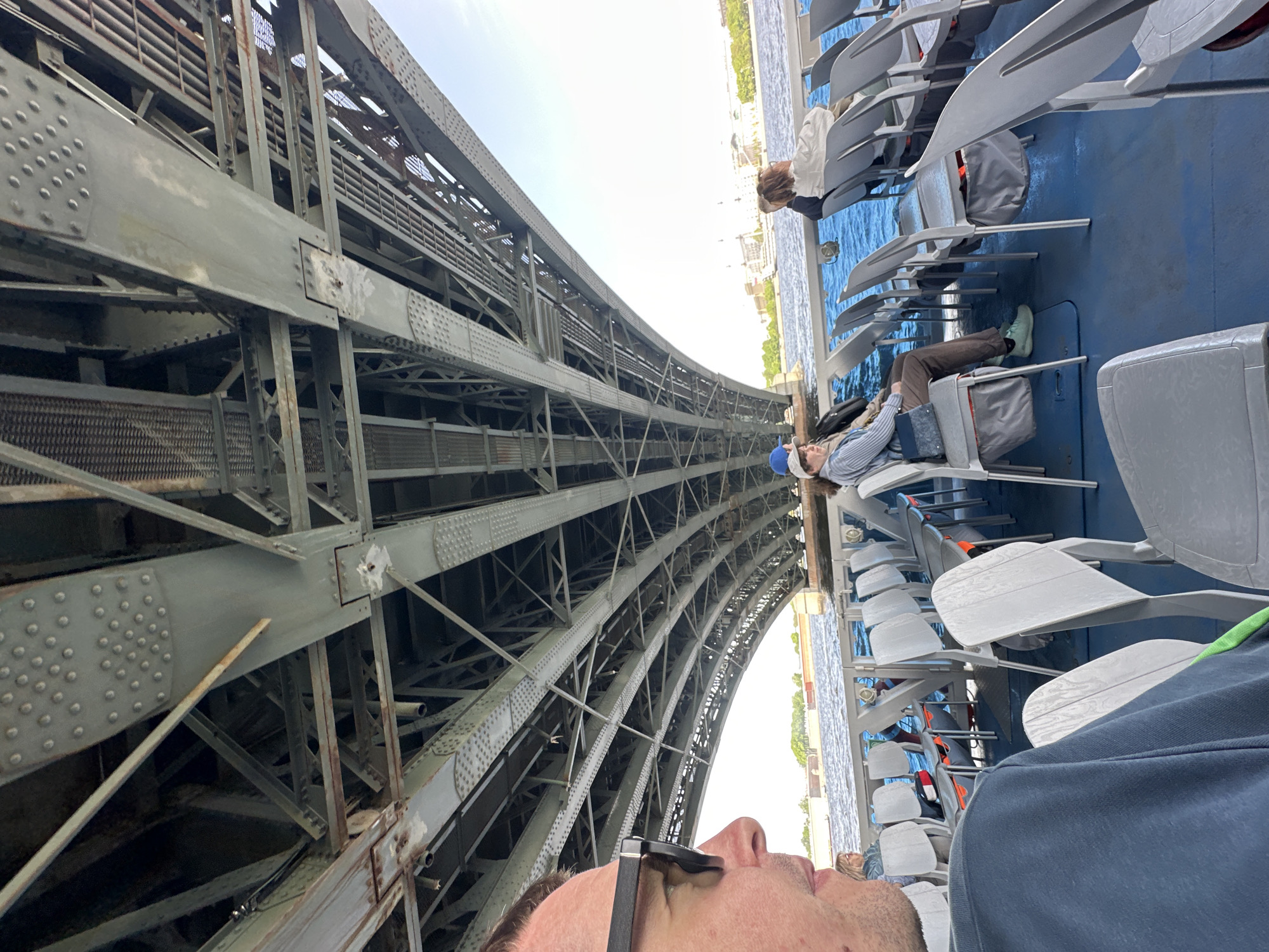 A low-angle, rotated shot depicts a gray steel bridge above a body of water. The bridge's architecture shows a latticework pattern of girders, plates, and rivets. On a boat underneath, people sit in gray plastic chairs. A person is visible in the foreground with the edge of a pair of glasses and a partial view of their nose. In the middle ground, a person lies reclined across two chairs. The background reveals a cityscape across the water. The setting is likely Saint Petersburg, Russia, with the bridge potentially being one of the city's historical structures.