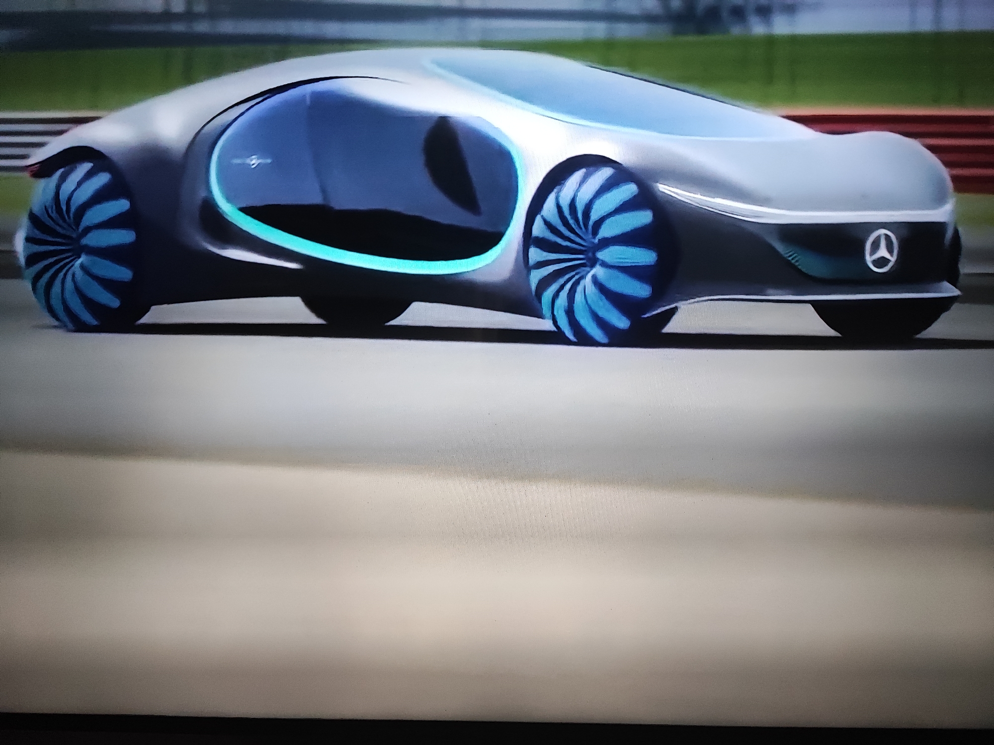 A digital rendering of a futuristic Mercedes-Benz concept car is shown in a close-up, side profile view. The car features a sleek, silver, aerodynamic body with large, circular windows that have a blue, glowing outline. The wheels are also a prominent design element, with a turbine-like structure and blue accents that appear to glow. The car is positioned on a flat, grey surface, resembling a road or track, with a blurred green background and red barrier in the distance, suggesting an outdoor setting, possibly a racetrack. The Mercedes-Benz logo is visible on the front of the car. The image appears to be a still from a video or a high-quality render, with a slightly grainy texture. The scene is static, with no people or actions depicted.