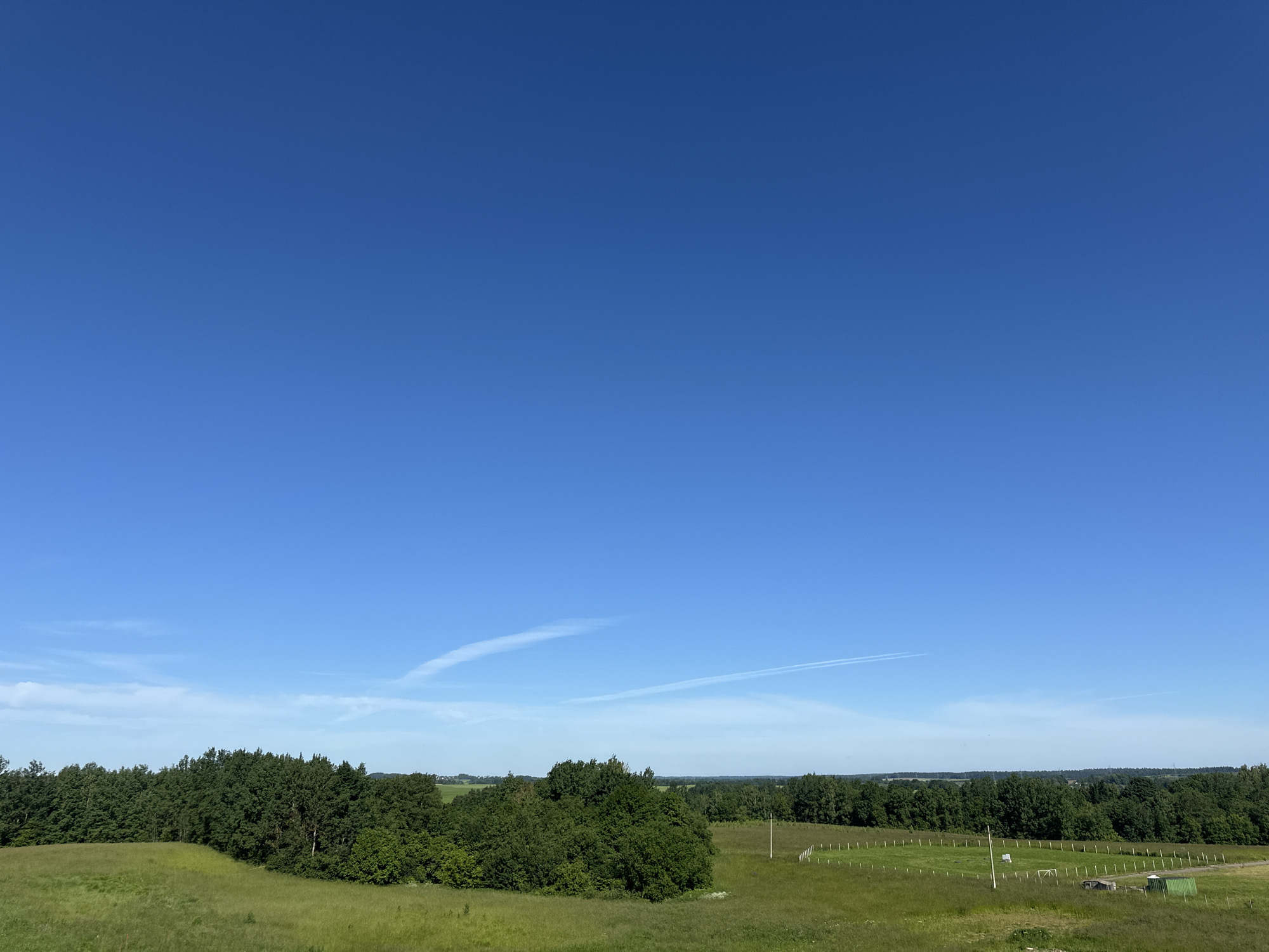 This is a wide, aerial landscape photograph taken on a clear, sunny day in the Шчомысліцкі сельскі Савет, Belarus. The top two-thirds of the image is dominated by a vibrant, cloudless blue sky. In the lower third, a rolling green landscape unfolds. A dense line of dark green deciduous trees forms a prominent barrier across the mid-ground. In front of the trees, to the left and center, is a meadow of tall, green grass. To the right of the trees, a fenced-off grassy area, possibly a sports field or paddock, is visible, marked by a white fence and several vertical posts. A single white pole with some equipment attached stands within this fenced area. Further in the distance, beyond the tree line, hints of more greenery and possibly some buildings can be seen. The overall scene depicts a rural, natural environment.