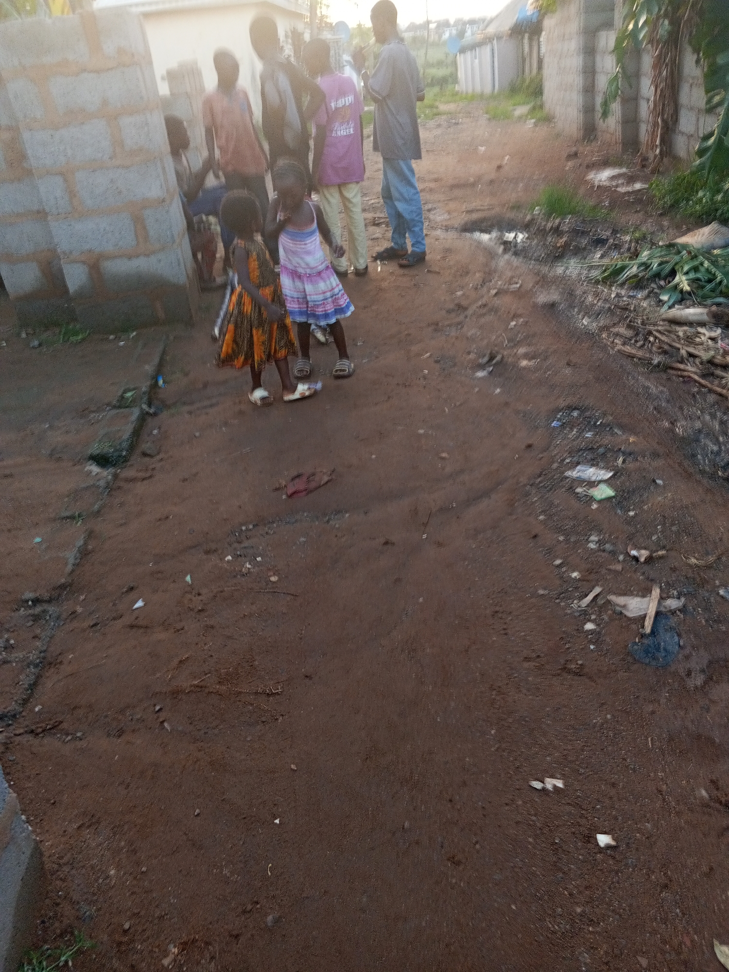 A group of people, including two young girls in dresses and sandals, are standing on a dirt road in what appears to be a residential area in Abuja, Nigeria. The road is uneven and has debris scattered on it. Several cinder-block buildings are visible. Some of the people are wearing casual clothing, and one person has on a purple shirt with text on the back. The light suggests it is either early morning or late afternoon.