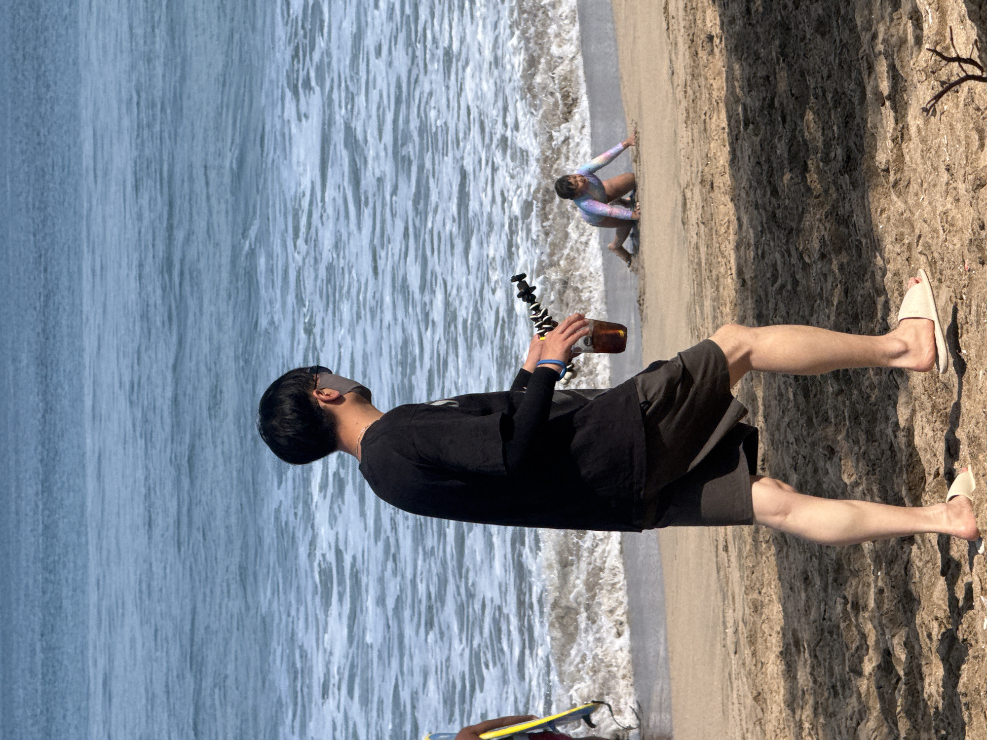 An adult male wearing a black t-shirt and black shorts walks on a sandy beach in Kuta, Indonesia, holding a drink in a plastic cup and a black tripod in his right hand. A child in a colorful swimsuit is crawling on the sand near the water's edge. The ocean is visible in the background with waves breaking on the shore. Another person holding a yellow surfboard is partially visible on the left side of the frame.