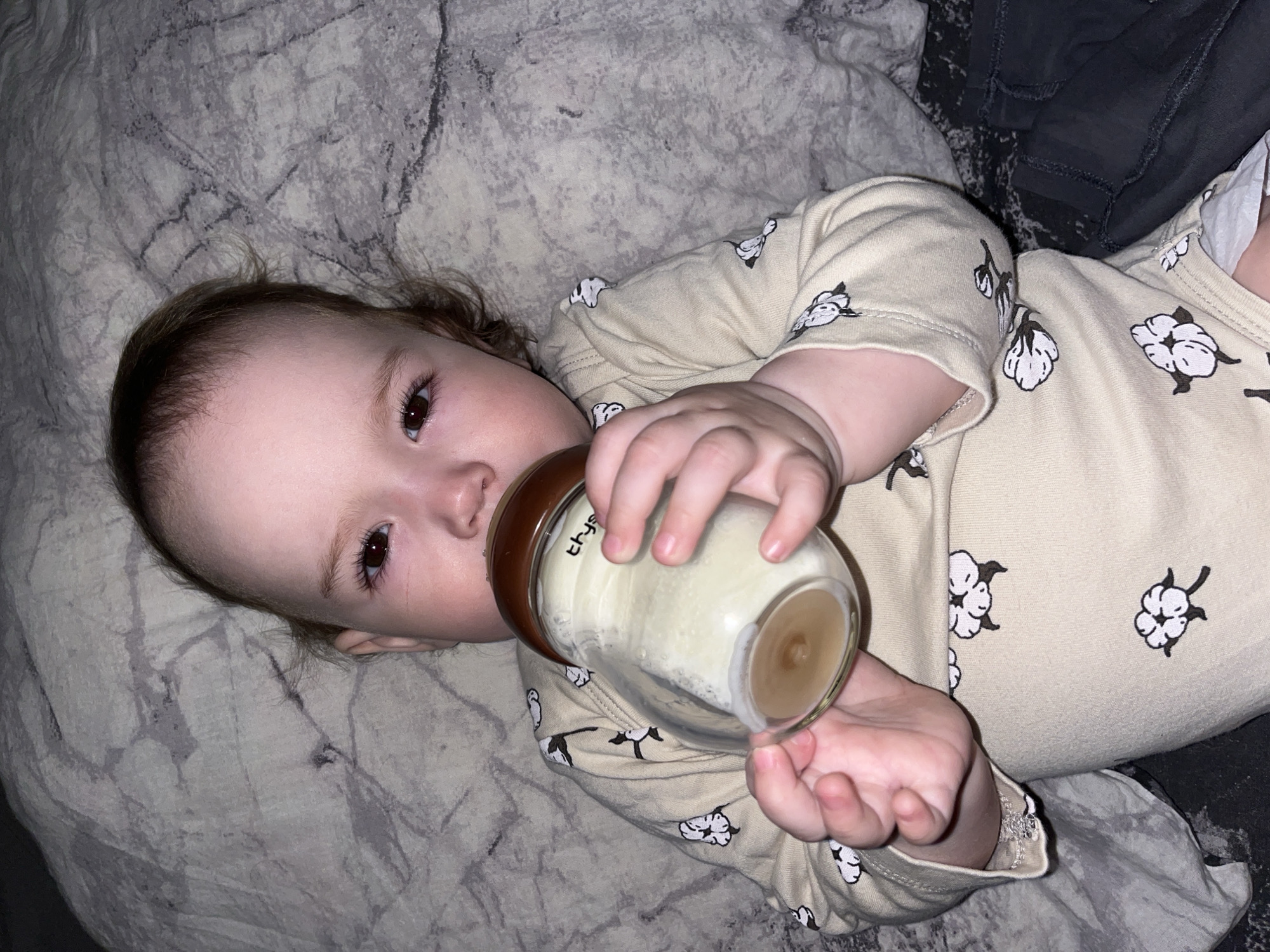 A baby lies on a patterned grey and white blanket and drinks from a baby bottle. The baby is wearing a light beige onesie with a repeating white cotton blossom pattern. The bottle is clear with a brown nipple and cap and contains a white liquid. The baby is positioned slightly off-center, with the head towards the left side of the image. Part of a dark grey garment is visible on the right side of the image. The location Shushary, Russia is not discernible from the image.