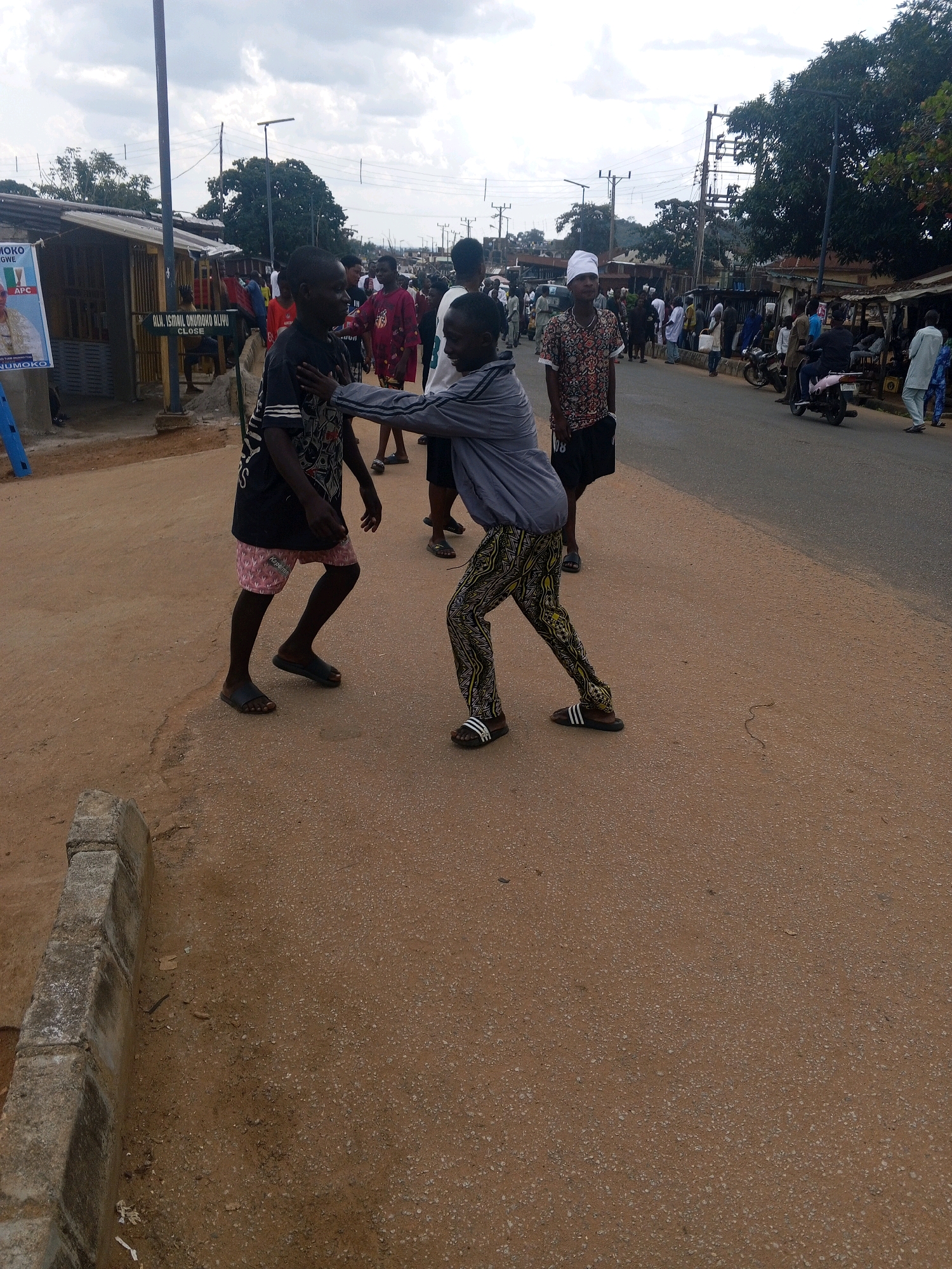 The image depicts a street scene in Okengwe, Nigeria, under a cloudy sky. In the foreground, two young boys are engaged in what appears to be a playful interaction, one wearing a black t-shirt with graphics and pink patterned shorts, the other in a grey athletic jacket and patterned trousers. They are standing on a dusty, light brown road with a concrete curb to the left. In the background, several people are gathered along the sides of the road, near market stalls and buildings. Some are standing and talking, while others are seated. There are also a few motorcycles and a small truck visible. A billboard with an image of a person and text is present on the left side of the image. The environment suggests a busy, communal area.