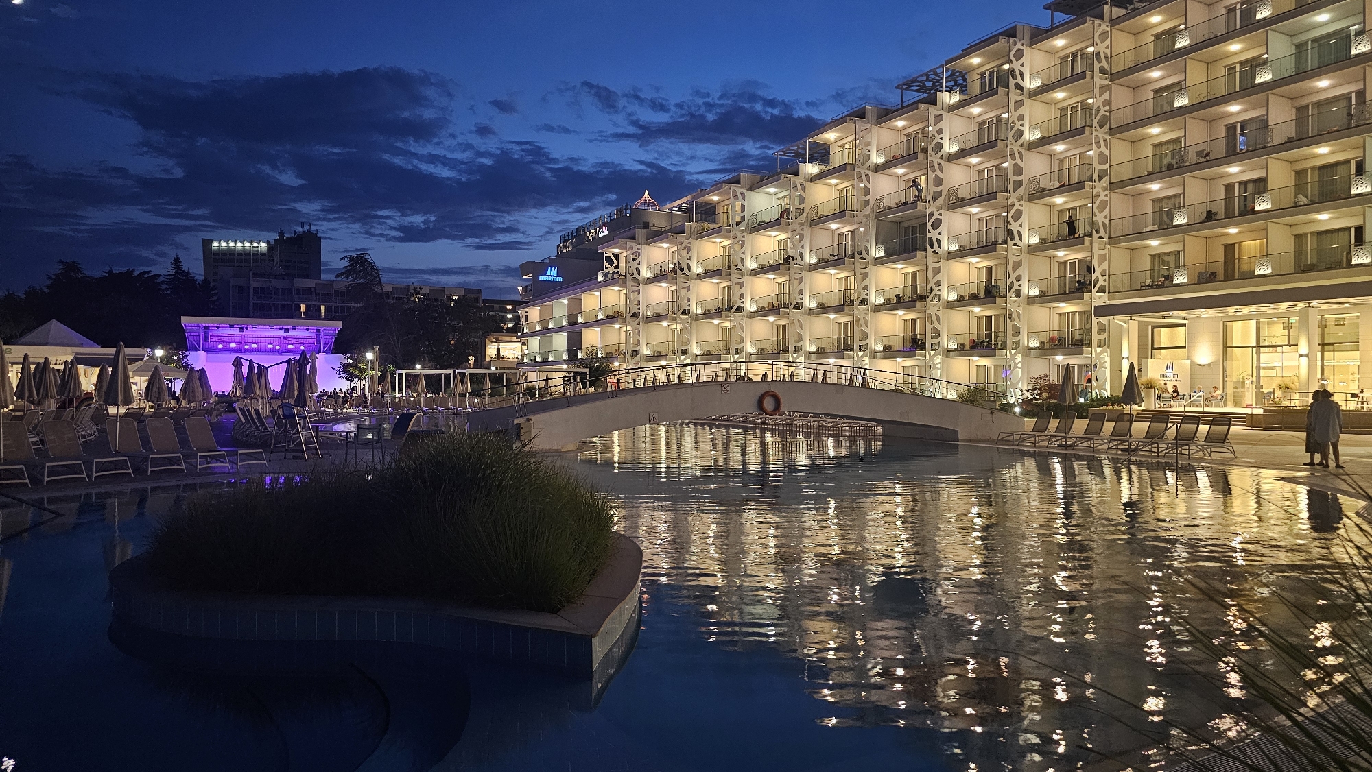 The image shows a resort in the city of Obrochishte, Bulgaria at dusk. A large, modern hotel with many lit windows and balconies dominates the right side of the frame. A pedestrian bridge arches over a body of water, reflecting the lights of the hotel and the twilight sky. In the foreground, a swimming pool is visible, with lounge chairs and umbrellas arranged around it. A stage with purple lighting is set up in the distance, with some people gathered. The sky is a deep blue, with scattered clouds. The overall scene conveys a sense of relaxation and evening entertainment.