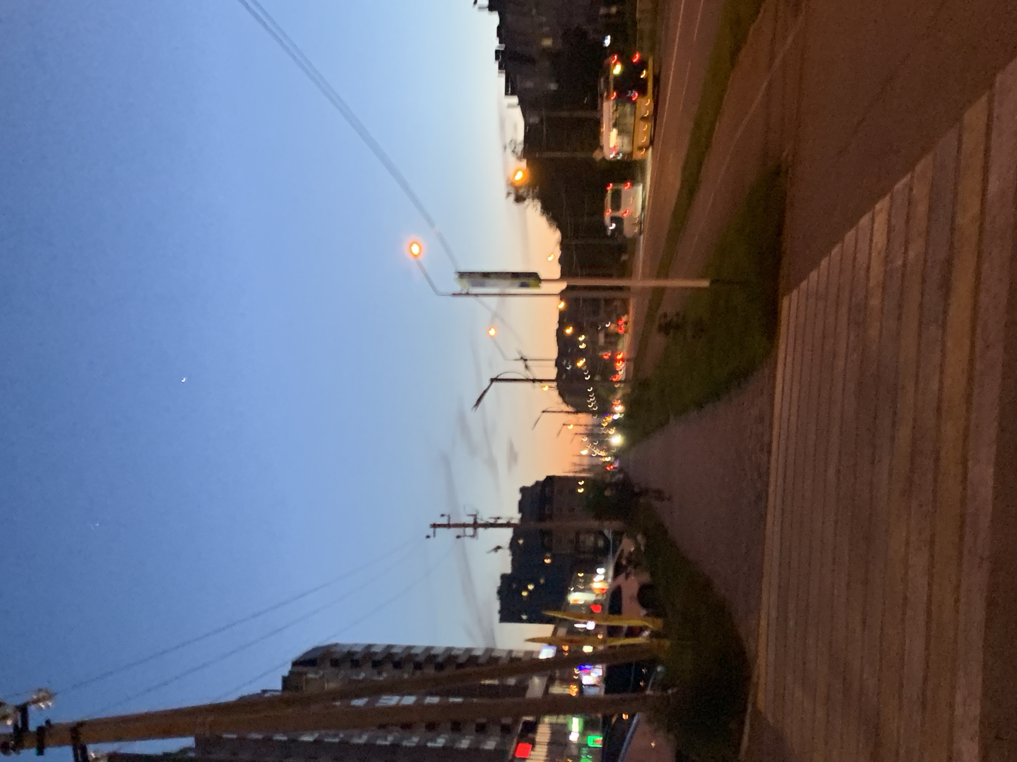 This image depicts a bustling urban street scene at dusk or early night in Stavropol, Russia. The sky, a clear expanse, transitions from a deep twilight blue overhead to a warm orange and yellow hue near the horizon, indicating the recent setting of the sun. A delicate crescent moon is subtly visible in the upper left of the sky.
The main subject is a city street, illuminated by numerous streetlights that cast an orange-yellow glow. Multi-story buildings, appearing as dark silhouettes against the twilight sky, line the left side of the street, with some interior lights and ground-floor signs faintly visible. Utility poles and overhead wires crisscross the sky, adding to the urban infrastructure.
The asphalt road carries traffic, with several vehicles visible. A yellow bus and another lighter-colored bus are discernable, their lights contributing to the blurred streaks of light from other cars in the distance. Traffic lights, showing red and yellow signals, are also part of the urban landscape. To the right, a wide pedestrian pathway or sidewalk, possibly a crosswalk due to its striped pattern, runs parallel to the road. Patches of green vegetation are visible along the roadside. The overall scene conveys the atmosphere of a city transitioning into night, with ongoing vehicle activity. No discernible text is visible on any signs or buildings.