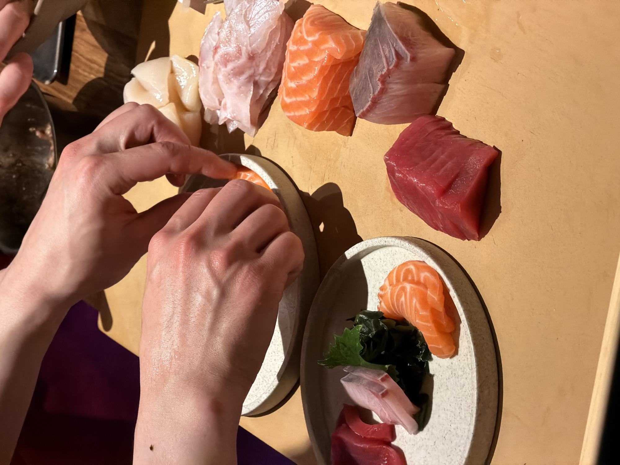 The image provides an overhead view of hands meticulously arranging various cuts of fresh raw fish, indicative of sashimi preparation, onto small, round ceramic plates. This activity occurs on a light brown wooden surface, likely a preparation counter or dining table in an indoor setting within Moscow, Russia.
In the upper part of the frame, several portions of fresh seafood are displayed: vibrant orange salmon, rich dark red tuna, creamy white scallops, and delicate light pink/white fish (possibly snapper or sea bream). A pair of hands is centrally positioned, carefully placing a piece of salmon onto one of the light-colored, speckled plates. To the lower right, a second plate is already prepared, showcasing an artful arrangement of salmon, tuna, a lighter-colored fish, and dark green seaweed.
The scene captures a precise and focused moment of food plating, highlighting the quality and presentation of the raw fish. The lighting suggests an indoor environment, with no discernible cues for the time of day or external weather. There is no visible text within the image.