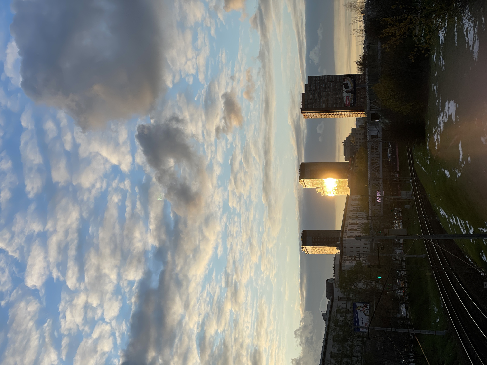 The image captures a vibrant golden hour scene over an urban landscape with prominent railway tracks in Khimki, Russia. The time of day is either sunrise or sunset, indicated by the warm, low-angled sunlight that casts intense reflections and highlights on the clouds and buildings.
The sky occupies a significant portion of the frame, filled with a diverse array of cumulus and stratocumulus clouds, illuminated from behind by the sun. Patches of clear, light blue sky are interspersed among the cloud formations, which range in color from soft white and gold where touched by sunlight, to darker grays in the shadows.
In the mid-ground, several tall, multi-story buildings are visible. The sun's bright disc is partially obscured by one of these structures, causing a brilliant flare and reflecting intensely off its facade. A large billboard on a prominent building on the right displays the text "ТОБАГО" with the slogan "МЕНЯЕТ ЖИЗНЬ НА ЛУЧШЕЕ" (Changes life for the better), accompanied by an image of a white car. Another, smaller billboard on the left, closer to the foreground, shows fragmented text including "опт.рынок" (wholesale market) and "Скидки до 40%".
The foreground features a railway system with multiple parallel tracks and associated overhead electrical lines supported by metal poles. Adjacent to the tracks on the right is a green grassy area dotted with scattered white patches, which appear to be snow or frost, suggesting cold weather conditions. Beyond the railway, a line of trees with varying degrees of foliage forms the horizon.
No human activity or people are visible in the image, which presents a static, contemplative view of an urban environment at a picturesque time of day, blending natural light with man-made structures and infrastructure.