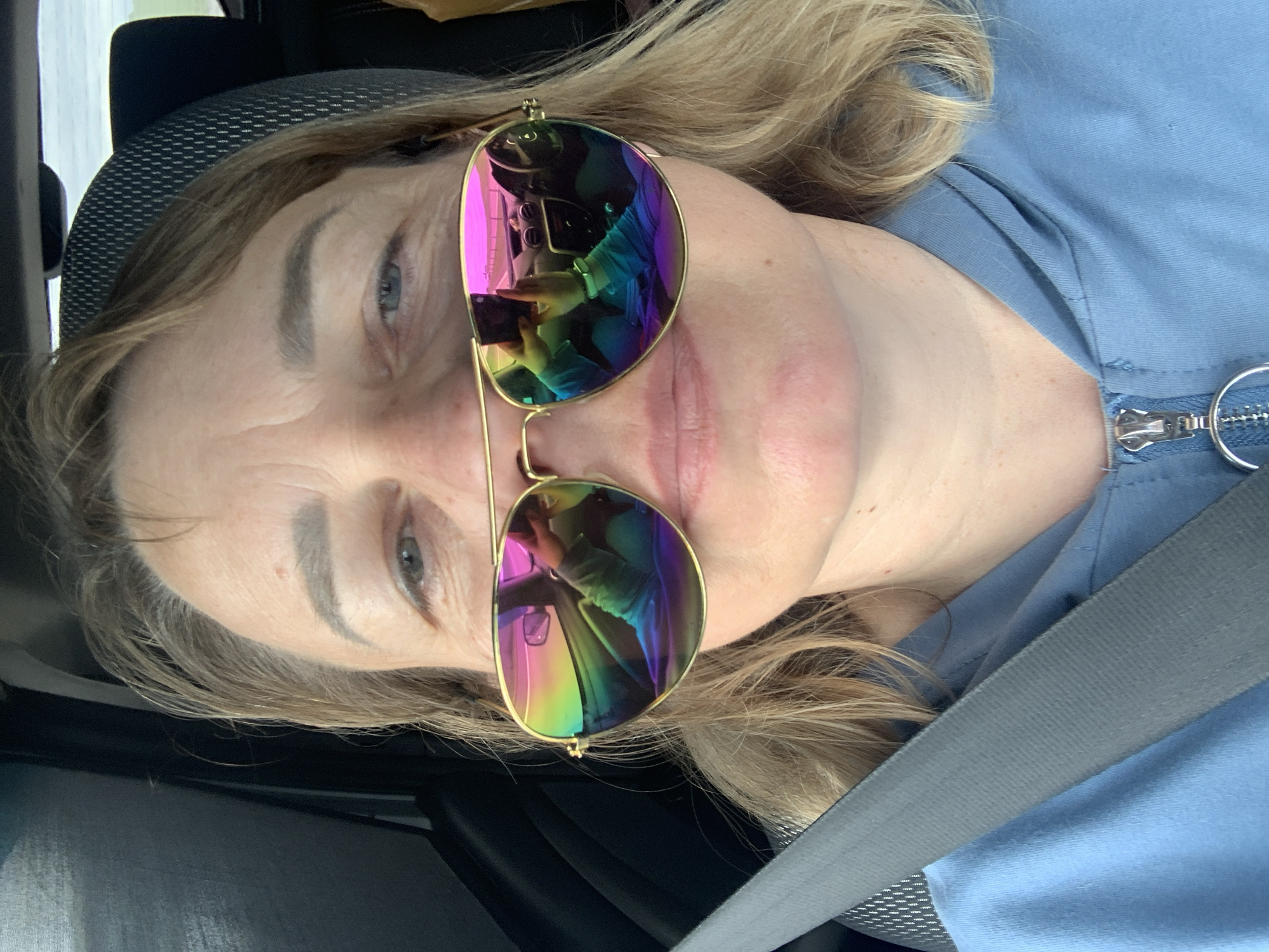 A close-up selfie shows a woman wearing sunglasses with rainbow-colored lenses. She is in the driver's seat of a car, with the car's dashboard and steering wheel reflected in her sunglasses. The woman is wearing a blue hoodie with the zipper partially down. A seatbelt is visible across her chest. The background is out of focus, but appears to be a road with some greenery. The scene is consistent with a person taking a selfie while driving in the Азерскі сельскі Савет region of Belarus.