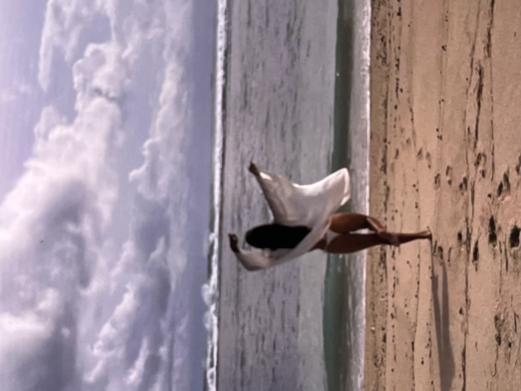 A woman lying on a surfboard on a beach in Unknown, Indonesia. The woman is wearing a white cover-up and a bikini. The beach is sandy and there are clouds in the sky. The ocean is in the background.