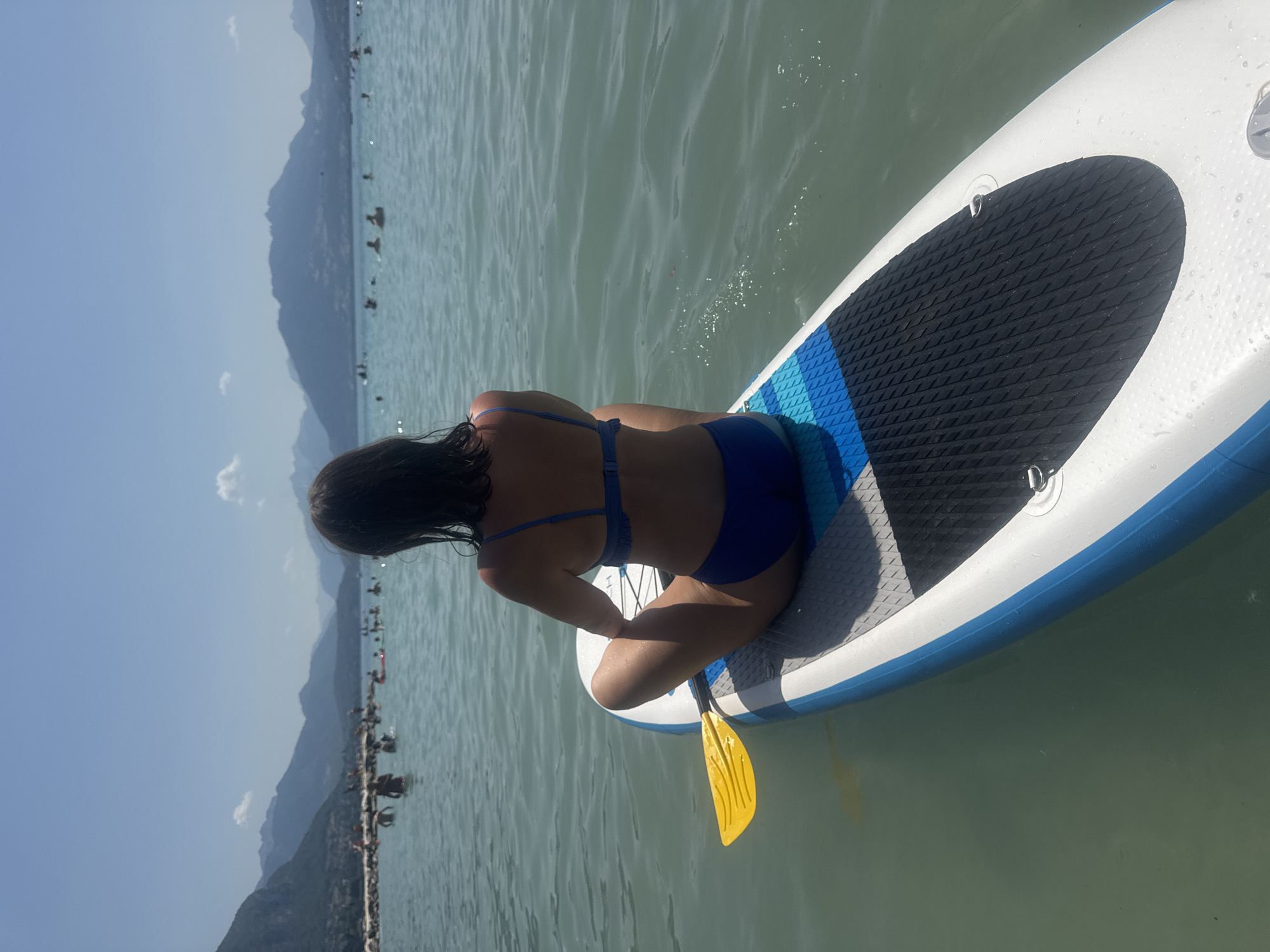A woman in a blue bikini sits on a paddleboard on Lake Annecy, France, facing away from the camera. The paddleboard, white with blue accents, has a yellow paddle resting on its side. In the background, the clear blue water is dotted with numerous people swimming and enjoying the lake. Beyond the lake, a shoreline with a rocky breakwater is visible, and in the distance, a range of majestic mountains under a clear blue sky completes the scenic view.