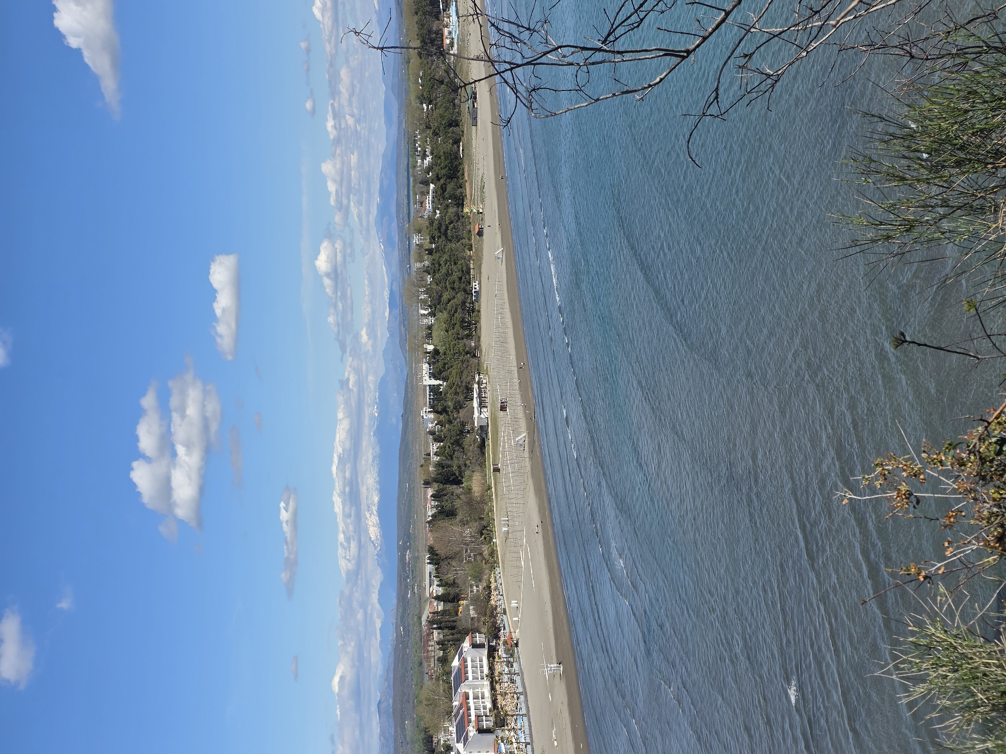 An elevated, long-shot shows a beach in Ulcinj - Ulqin, Montenegro. The Ionian Sea is to the right. The sand stretches along the coast; many white canopies line the beach and trees and buildings are located behind them. Further, the mountains are in the background. The sky on the left is blue with small, puffy clouds. A tree and a green bush grow in the immediate foreground.