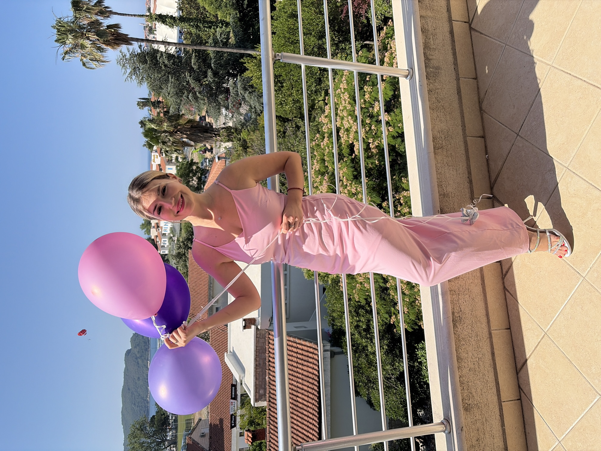 A blonde woman in a pink slip dress and jeweled sandals stands on a tiled balcony in Budva, Montenegro, holding three balloons, two purple and one pink, on a sunny day. The woman is smiling at the camera. Behind her, a railing separates the balcony from a lush green area with trees and buildings with red tiled roofs. Palm trees and more buildings are visible in the background, along with a clear blue sky. A paraglider can be seen in the distance.