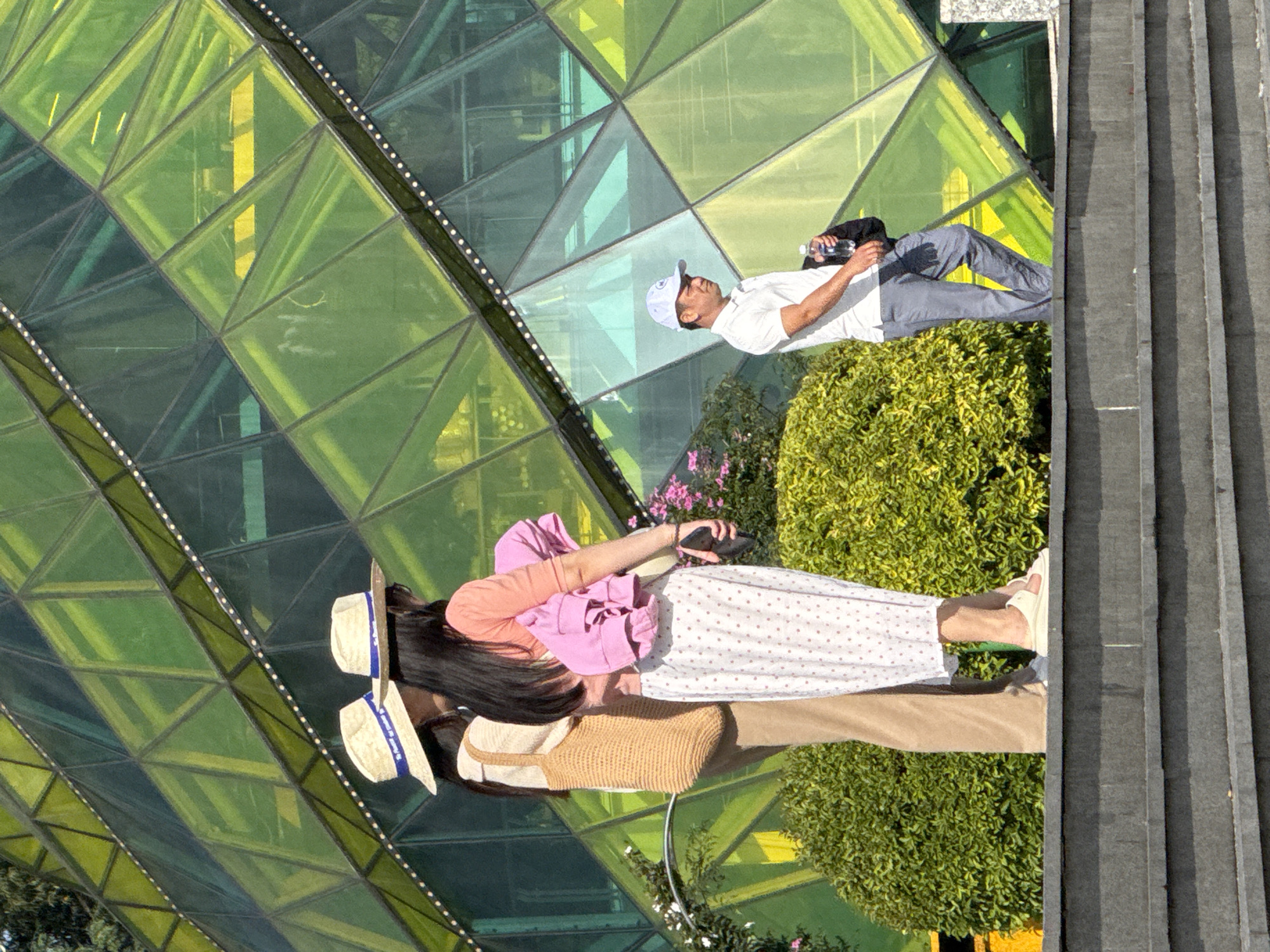 The image shows a scene in front of the Da Lat Railway Station in Da Lat, Vietnam. The station's distinctive architecture, featuring green and yellow geometric glass panels, is visible in the background.
In the foreground, two individuals, appearing to be women, stand facing a building. Both are wearing straw hats. The woman on the left wears a beige top and light brown pants, carrying a woven straw bag. The woman on the right wears a pink short-sleeved top and a white skirt with pink polka dots, and is carrying a pink garment over her shoulder.
To the right, a man wearing a white polo shirt, grey pants, and a white baseball cap walks away from the camera, holding a plastic water bottle and a dark jacket. He is positioned on a lower level, separated by a set of dark grey stone steps.
The environment includes lush green bushes in front of the building. The lighting suggests it is daytime, with strong sunlight casting shadows. The overall scene captures a moment of people in an urban setting with a notable architectural landmark.