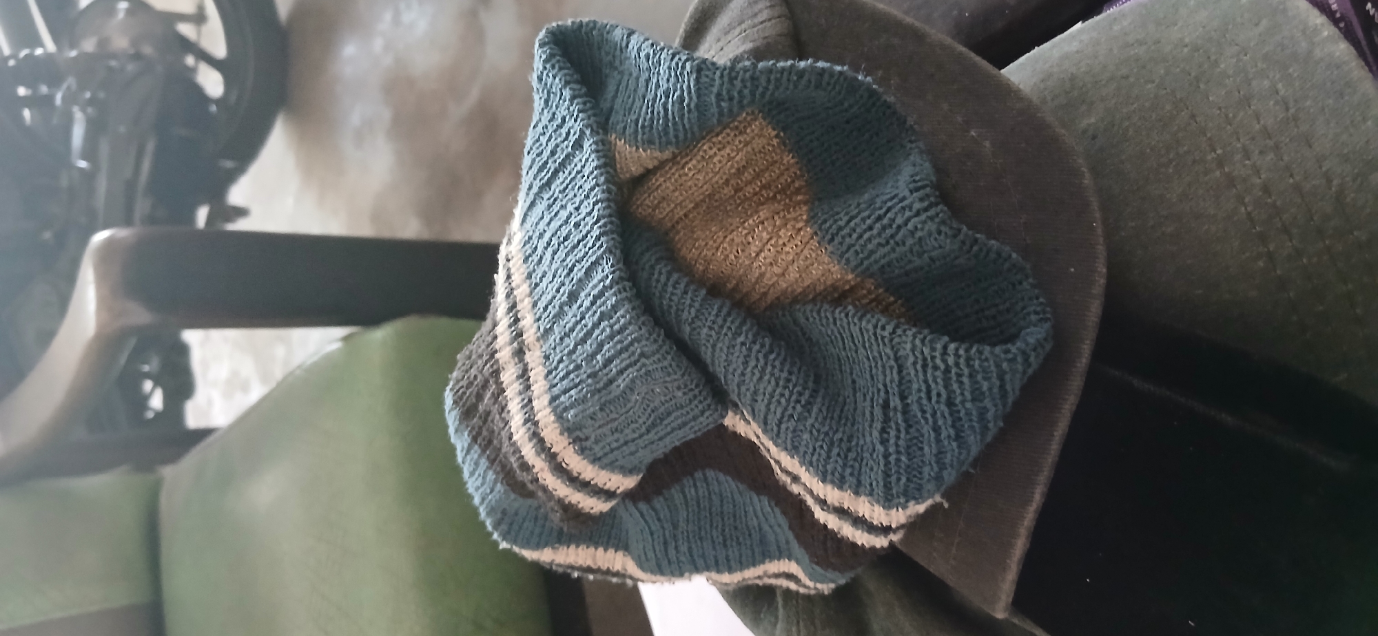 In Kademangan Kulon, Indonesia, a knitted sock with blue, white, and grey stripes is draped over a dark baseball cap. The sock is folded and bunched, revealing its textured weave. The cap appears to be made of a dark fabric, possibly cotton or a similar material. The scene is set indoors, with a blurred background that includes a green upholstered chair and the wheel of a motorcycle. The lighting suggests an indoor environment, possibly a room or a covered outdoor area.