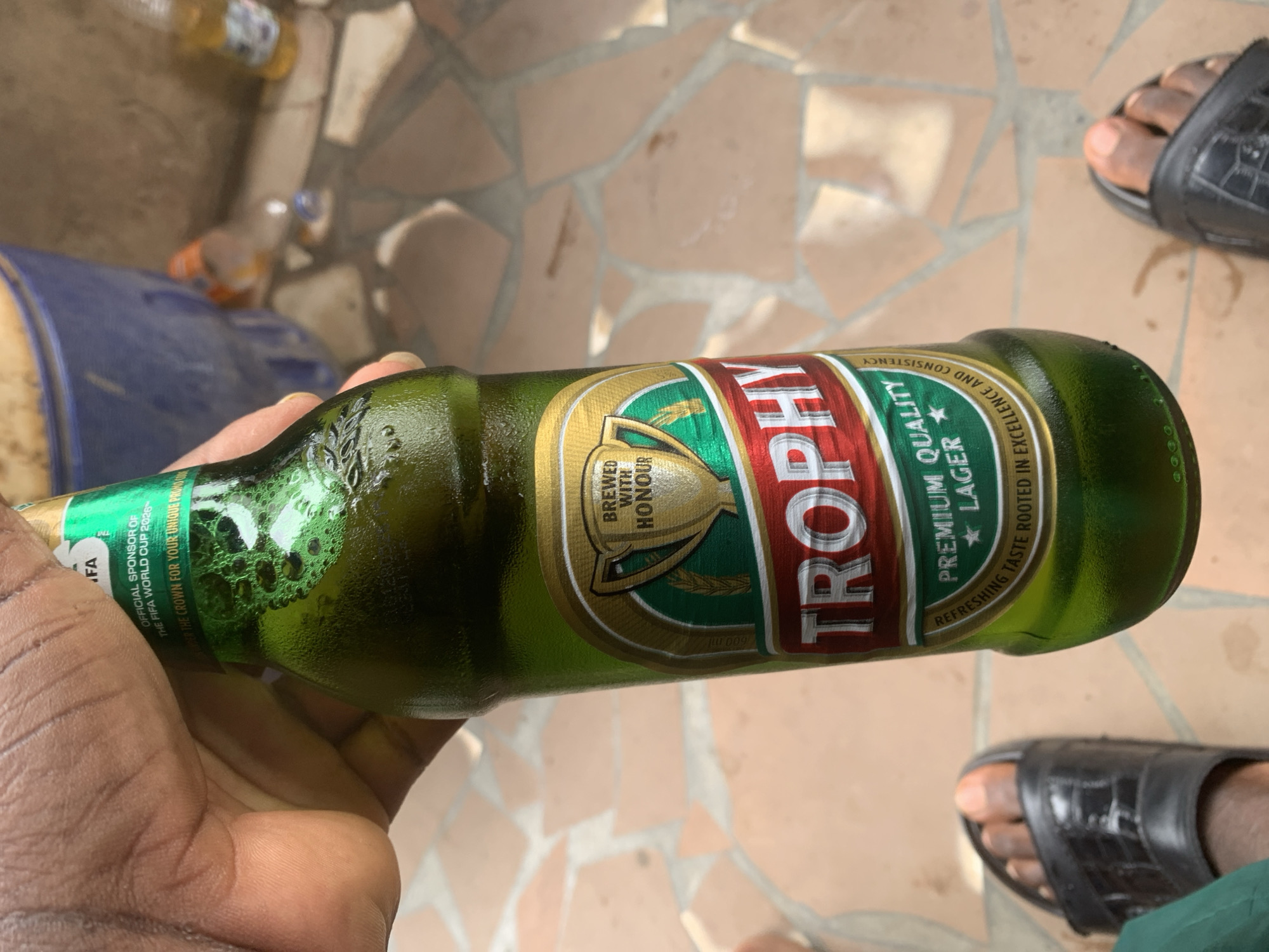 A person's hand holds a frosty green bottle of Trophy lager, a prominent Nigerian beer. The label, featuring a gold trophy and the words "Brewed With Honour" and "Trophy Premium Quality Lager," is clearly visible. The bottle appears to be sweating, indicating it is cold. The background shows a tiled patio, with a blue bucket and other discarded bottles suggesting an outdoor gathering. Two feet, clad in black open-toed sandals, are visible at the bottom of the frame, grounding the scene in a typical Nigerian setting, possibly in Shagamu.