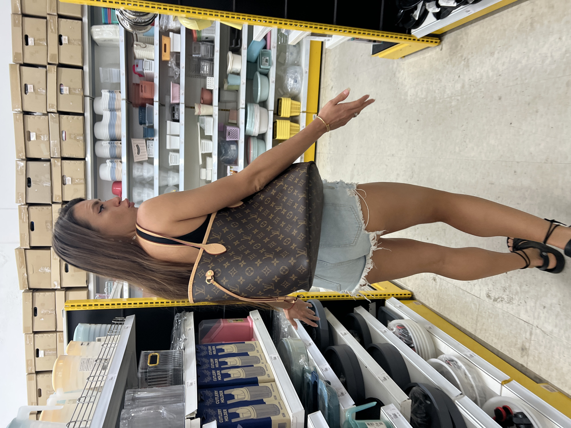 A woman with long brown hair and a Louis Vuitton tote bag is shopping in a store that sells housewares, possibly in Baan Lamai, Thailand. She is wearing a black tank top and denim shorts. The store has white shelves filled with various containers, baskets, and kitchenware. The woman is reaching out with her right hand towards a shelf.