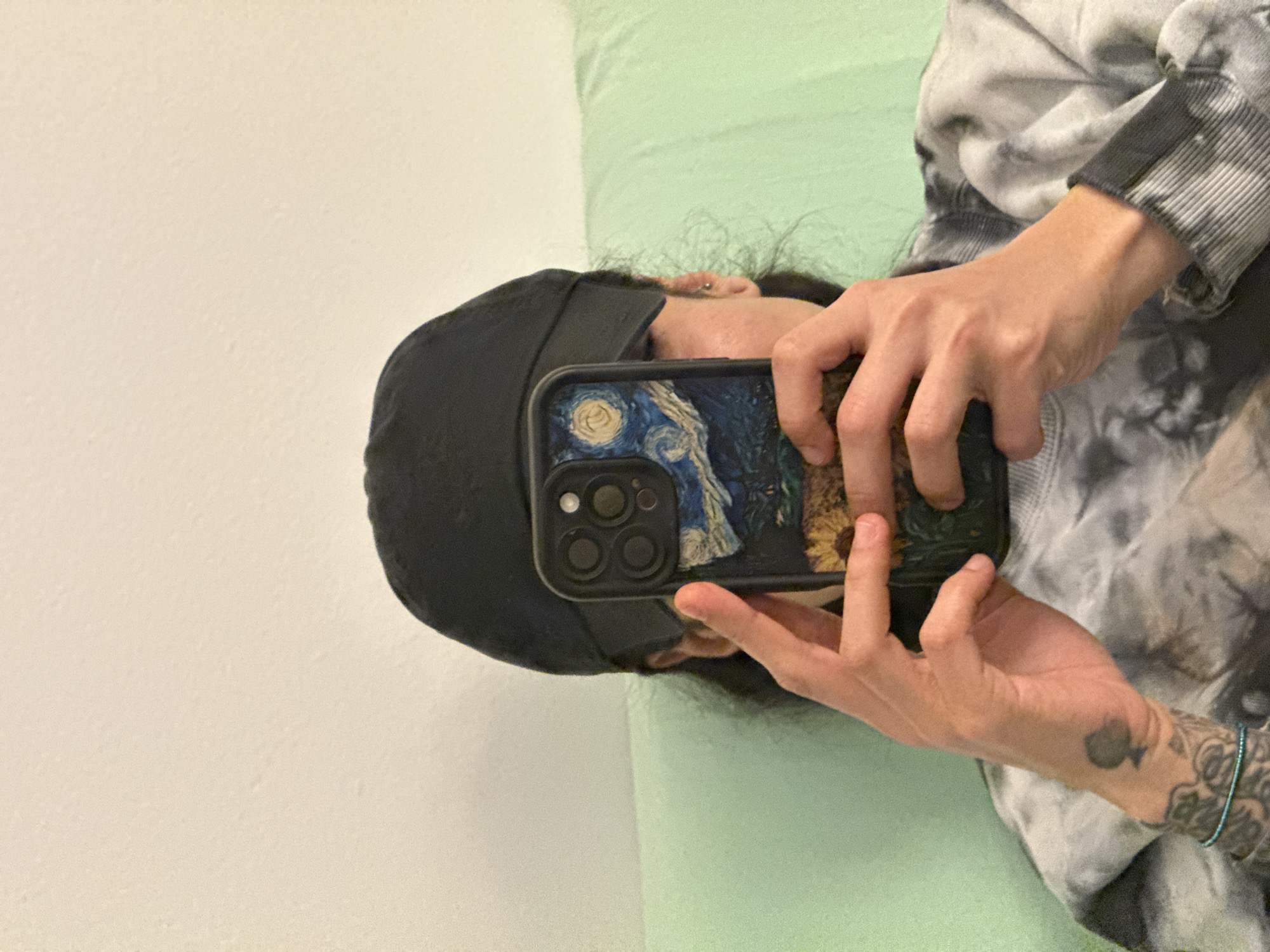 A person is taking a selfie with a smartphone featuring a case with artwork reminiscent of Vincent van Gogh's "Starry Night" and sunflowers. The person wears a dark-colored baseball cap, and a gray and white tie-dye patterned sweatshirt. They are sitting in front of a wall with a light green section, suggesting a cushion or headboard. The background features a white wall. There is no visible information to place this image in Barcelona, Spain.