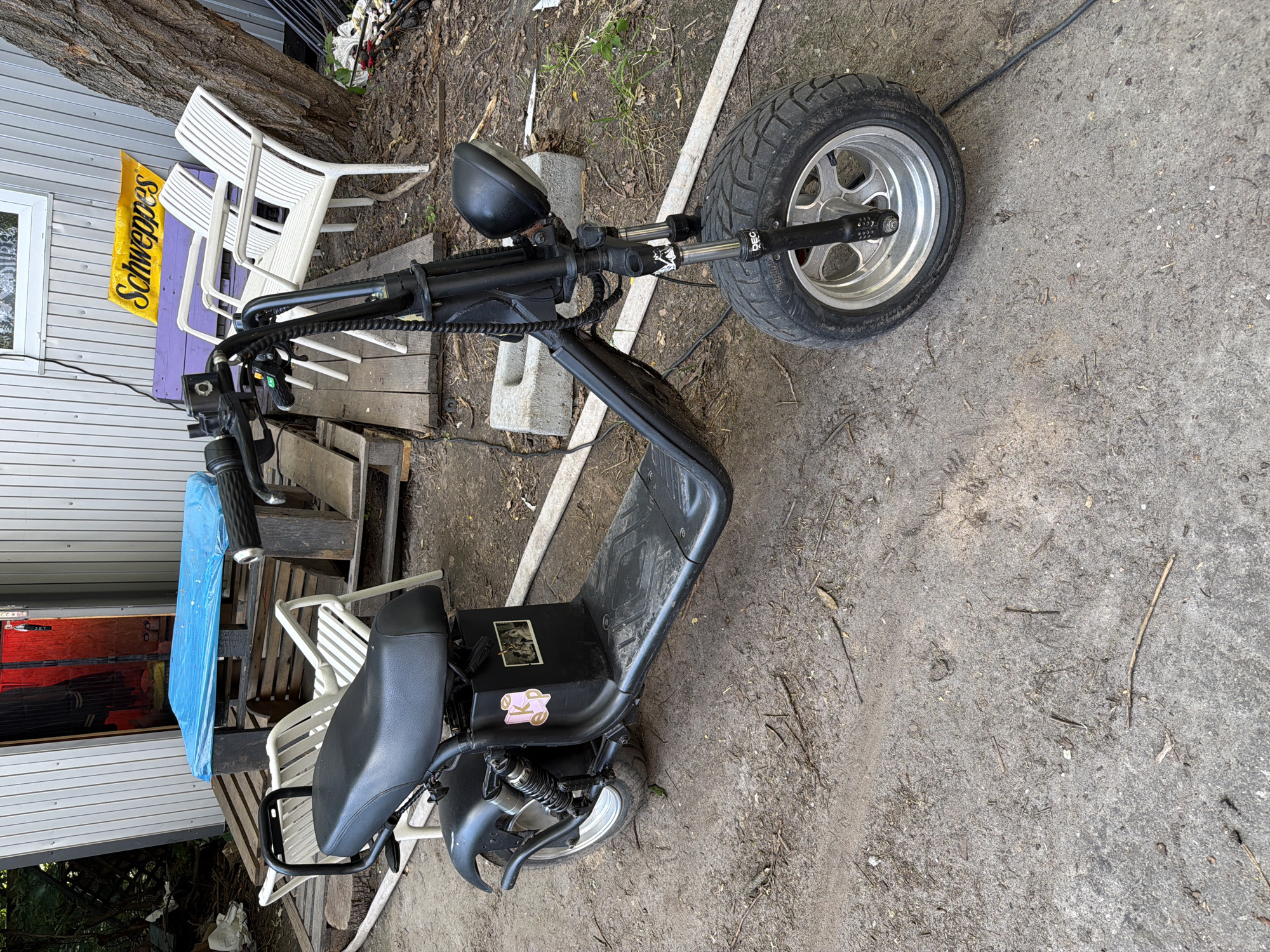 The image depicts a black electric scooter parked outdoors in what appears to be a somewhat unkempt urban or suburban setting. The scooter is positioned on a dirt and gravel surface, with scattered twigs and debris. In the background, a light-colored building with corrugated siding is visible, along with wooden pallets stacked near an open doorway revealing a red interior. Several white plastic chairs and a purple bench are present near the building. A bright yellow sign with the Schweppes logo is affixed to the wall. A large tree trunk is on the right side of the image. The scooter itself has a wide seat, thick tires, a single headlight, and handlebars with control levers. A sticker, partially obscured, is on the side panel of the scooter. The overall impression is one of an outdoor storage area or a casual backyard space. The location is stated to be in Kyiv, Ukraine.