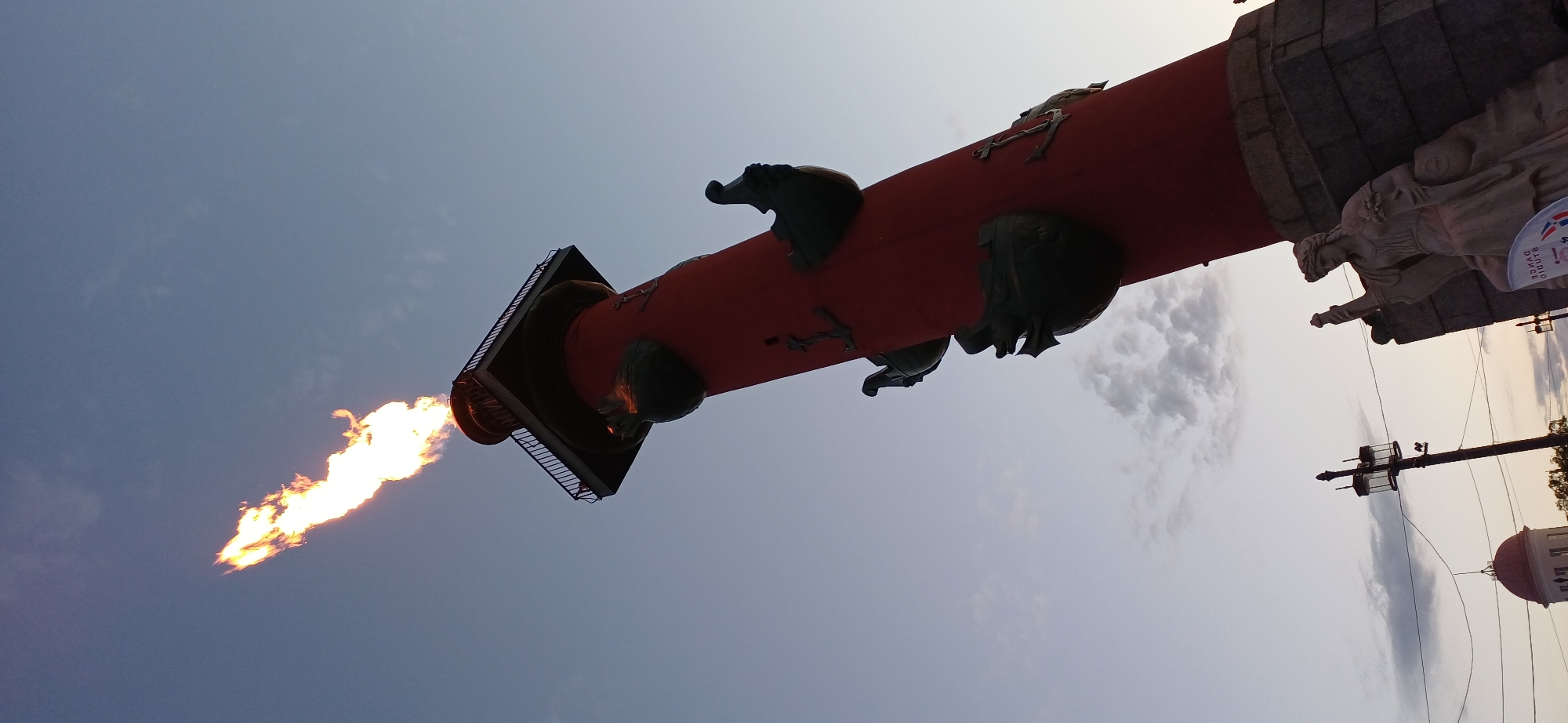 The image captures the Rostral Column, a prominent landmark in Saint Petersburg, Russia, situated on the Spit of Vasilievsky Island. A bright orange flame erupts from the top of the red, cylindrical column, which is adorned with anchors and sculptures of ships' prows. The column is angled upwards against a pale blue sky with scattered clouds. At the base of the column, a stone pedestal is visible, supporting a classical statue of a woman holding a lyre. In the background, to the left, a street lamp and the dome of a building with a red roof can be seen. The overall lighting suggests either dawn or dusk, with a soft, diffused light.