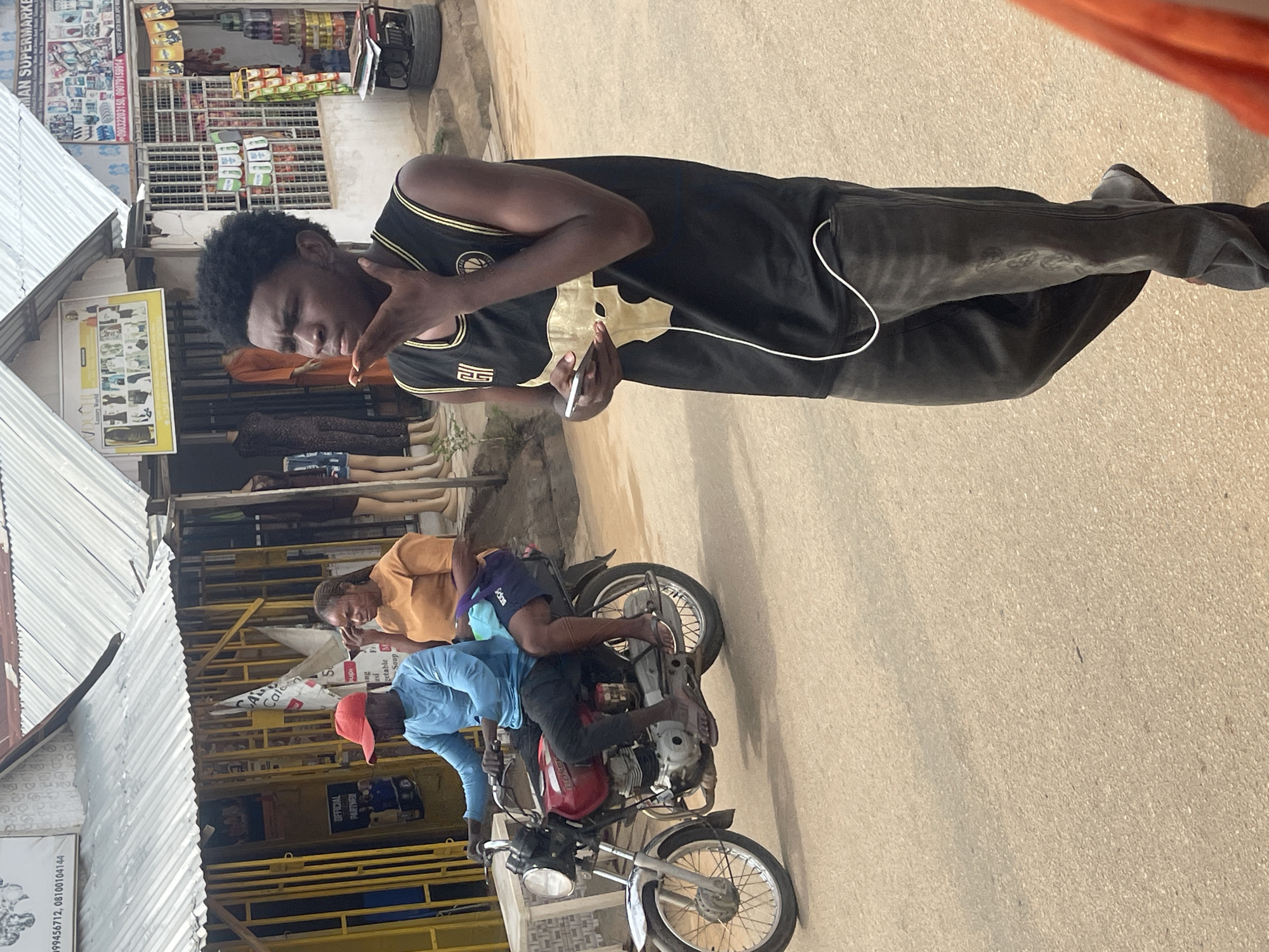 This image depicts a bustling street scene in Abuja, Nigeria, under bright daylight, likely mid-morning or afternoon, given the clear, direct sunlight.
In the foreground, a dark-skinned man with black curly hair is the primary subject. He wears a black sleeveless basketball-style jersey with gold accents, featuring what appears to be the Golden State Warriors logo on the chest and the number '25' on the lower right. He is holding a silver smartphone in his right hand, with a white cable (likely for earphones or charging) extending from it. His left hand is raised in a gesture, and his expression appears focused or slightly strained. He is also wearing dark trousers.
Behind him, along what seems to be a commercial street, are various shops with corrugated metal roofs. Visible signage includes "MAN SUPERMARKET" and fragments of other text such as "OFFICIAL PARTNER," "Caten 5," "getable Soup," and phone numbers: "#99456712, 08100104144". Mannequins displaying clothes are visible within the shop windows, suggesting clothing or general merchandise stores.
Further down the street, a red motorcycle is either parked or moving slowly. A man in a light blue long-sleeved shirt and a red baseball cap is driving, with his back mostly turned to the camera. Behind him, a dark-skinned woman with braided hair, wearing an orange-brown top and purple shorts, rides as a passenger. The ground is a light-colored paved surface.