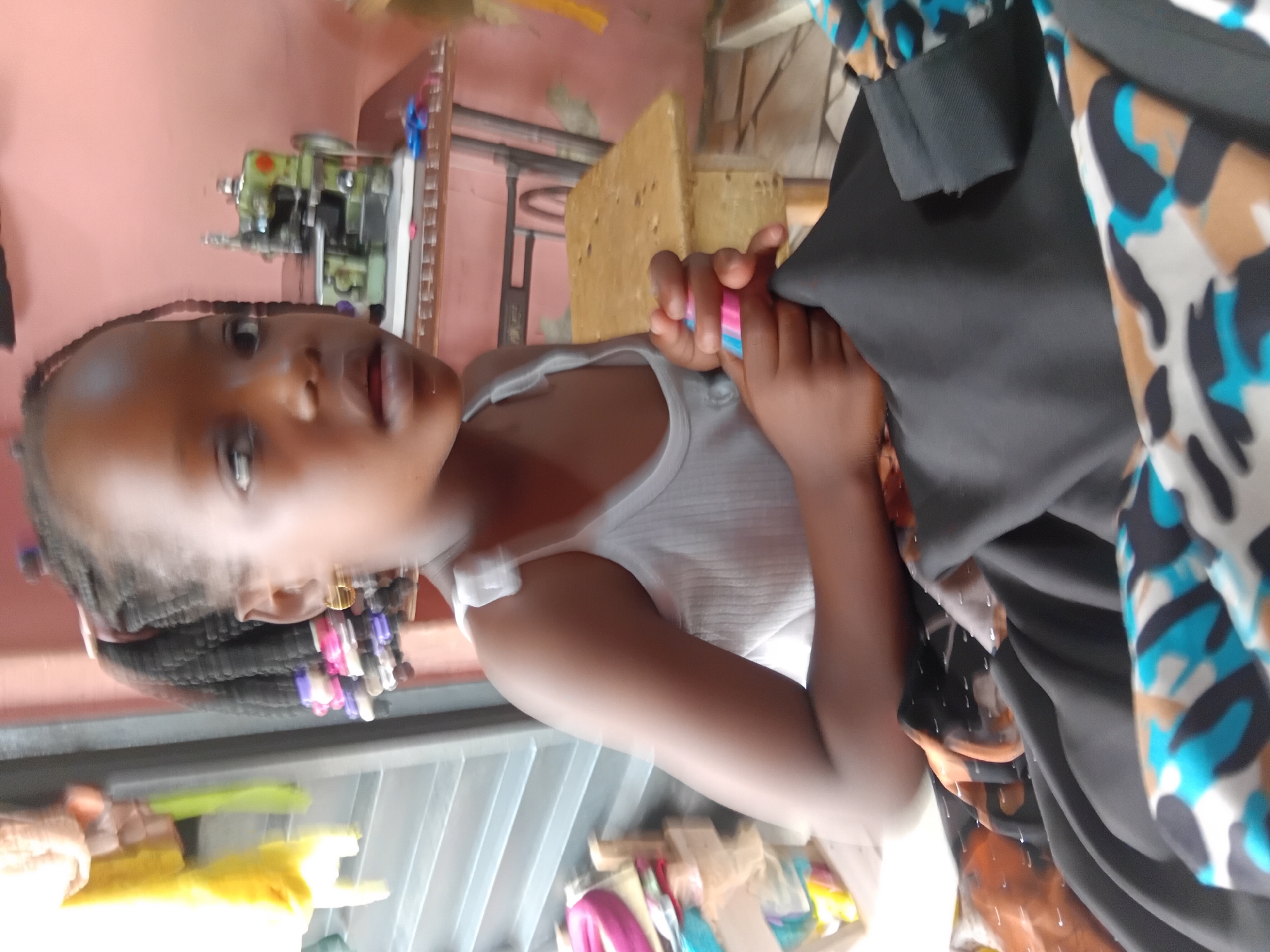 A young girl with braided hair adorns large gold hoop earrings. She is wearing a white tank top and holds a small object with a pink and blue striped pattern in her right hand. She is looking off to the side with her mouth slightly open. In the background, a vintage sewing machine sits on a table. The scene appears to be indoors, possibly a workshop or home.