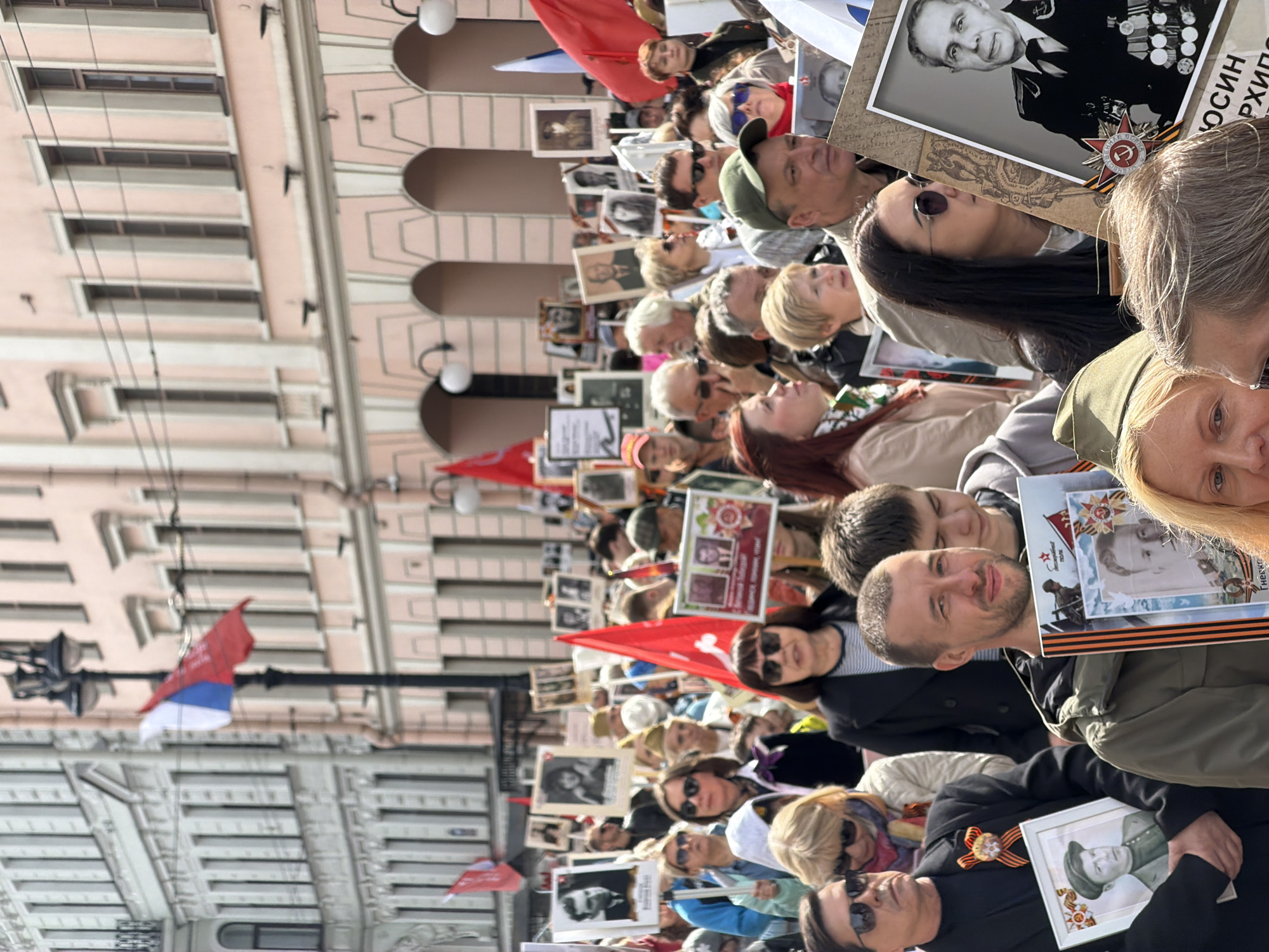 The image depicts a large crowd of people in Saint Petersburg, Russia, likely participating in a commemorative event like the "Immortal Regiment" march. Many individuals are holding portraits of deceased family members, presumably veterans of past wars. Red flags and Russian tricolor flags are visible. The environment appears to be a city street lined with buildings that are tall with light-colored facades. The weather is sunny.