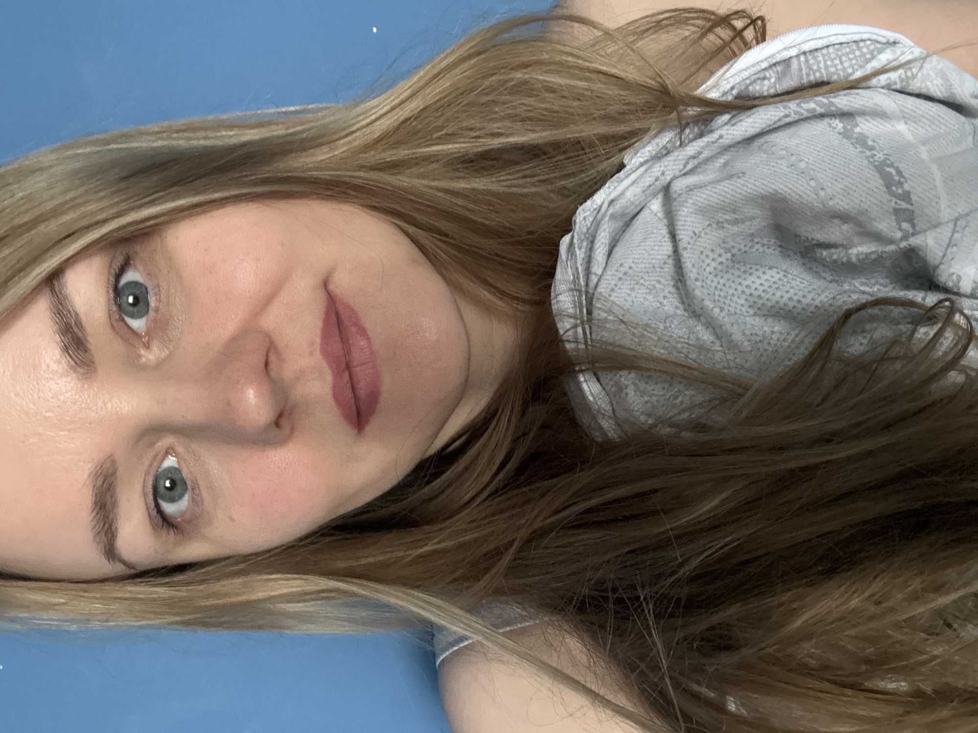A close-up shot shows a woman with light-colored eyes and long, light brown and blonde hair. She is wearing a light gray patterned top. The background is a solid blue. The location is unknown and cannot be determined from the image. There is no indication of the city of Chaikovsky, Russia.