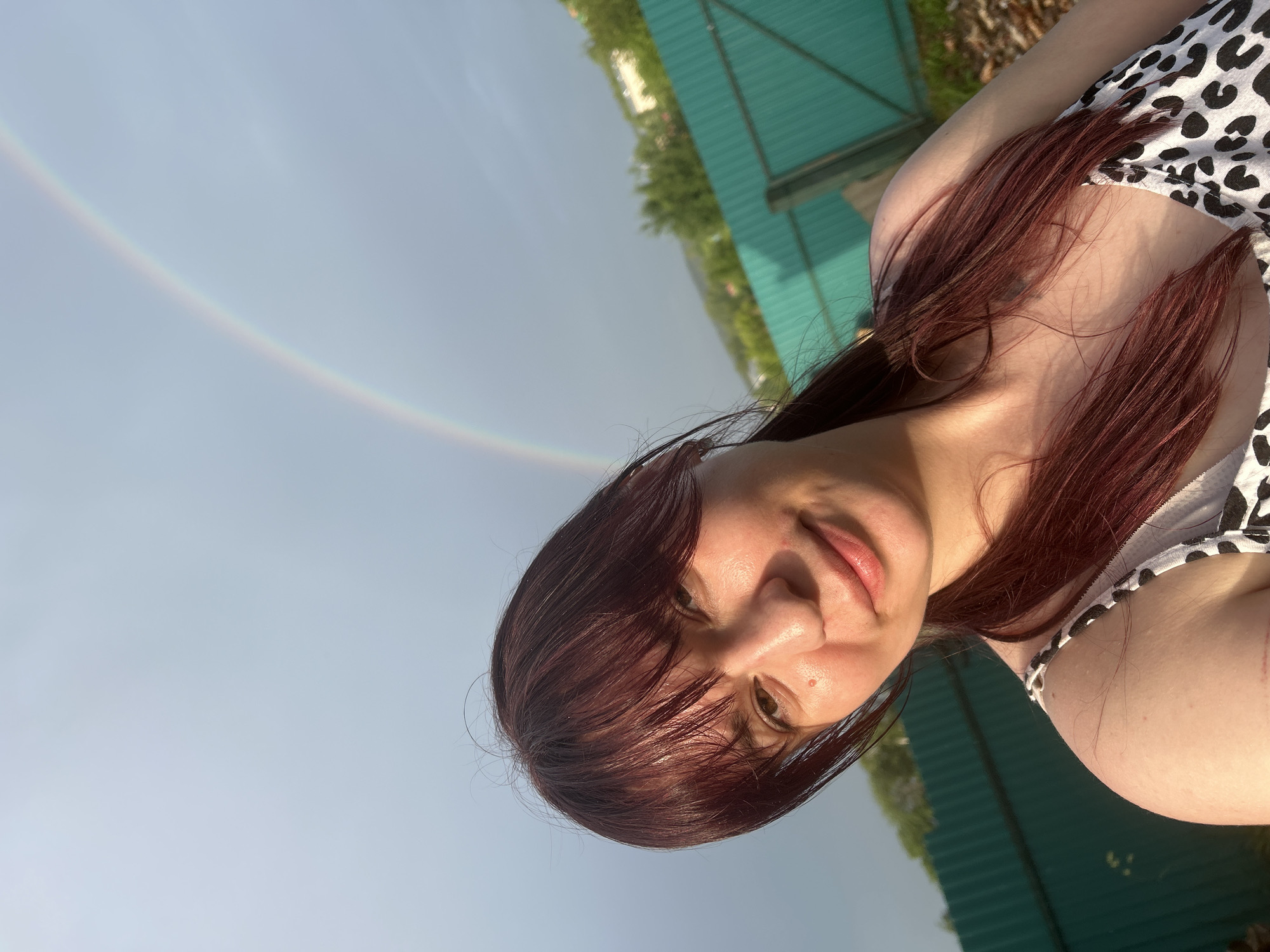 A medium shot captures a young woman with auburn hair, styled with bangs and pulled back into pigtails, facing the camera with a gentle smile. She is wearing a white dress with a black leopard print. Her skin appears slightly sun-kissed, with a mole visible on her cheek. Behind her, a faint rainbow arcs across a cloudy sky, suggesting recent rain. The background features a bright green corrugated metal fence, trees, and distant buildings, hinting at a suburban or rural setting. The lighting suggests it is late afternoon or early evening, with the sun casting a warm glow. The overall impression is of a casual, outdoor portrait taken after a rain shower. The specific location is the city of Fokino, Russia.