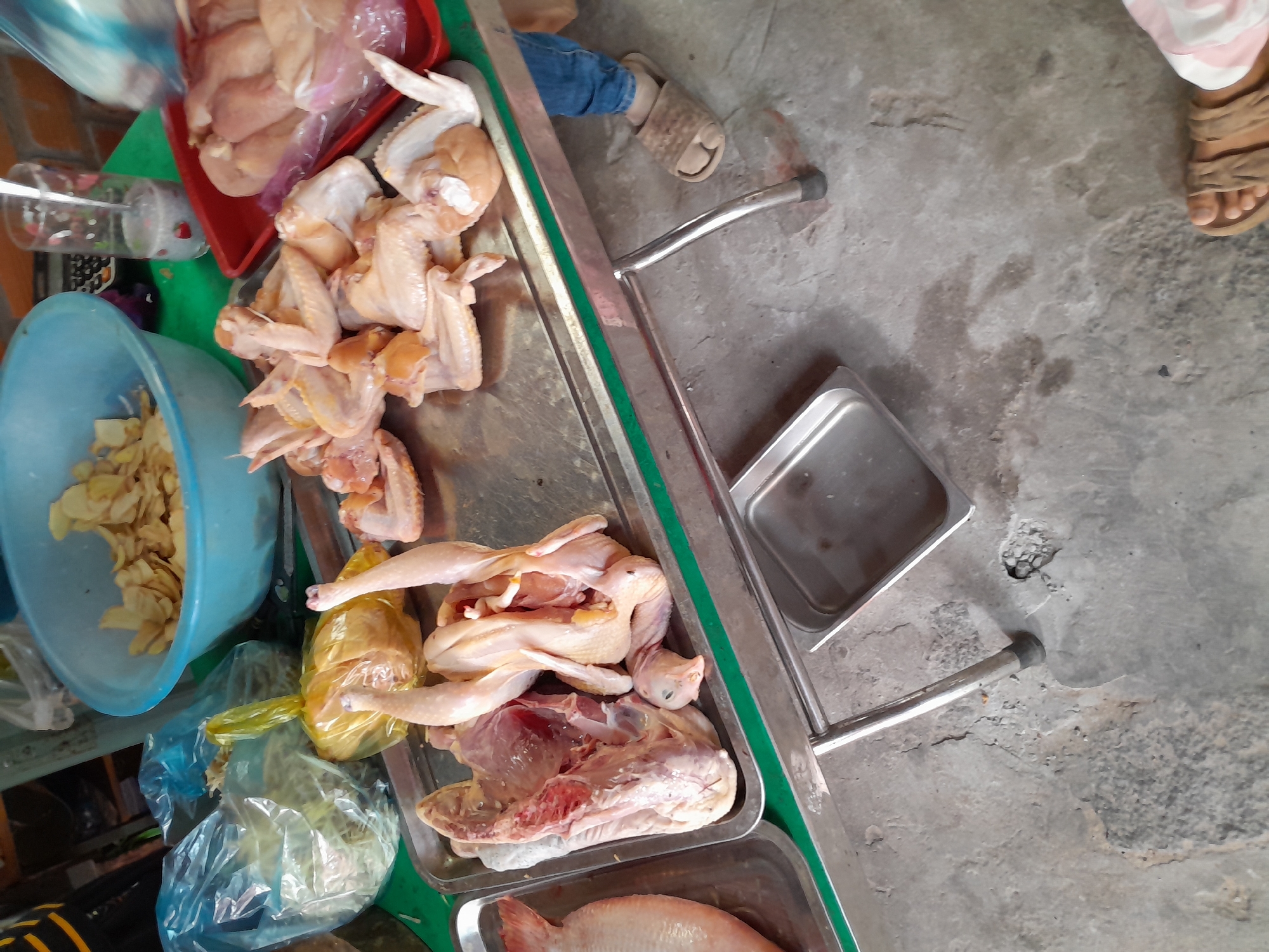 An overhead, slightly angled medium shot captures a market stall in Phu Thuy Ward, Vietnam, showcasing a selection of raw poultry and fish. The scene is dominated by a metal table displaying various trays and bowls of food items. In the foreground, a large, whole fish lies on the left side of a silver tray. To its right, on the same tray, are two whole, raw chickens, one appearing to be partially deboned, with its head and neck still attached. Another tray, positioned behind the first, holds a pile of raw chicken wings. Further back, a red plastic tray contains more raw chicken pieces, partially obscured by plastic packaging. To the left of the chicken wings, a blue plastic bowl is filled with thinly sliced, light-colored food items, possibly dried fruits or vegetables. Behind this bowl, a clear glass with a straw contains a light-colored liquid. The table itself has a green border. In the lower right of the frame, a person's legs are visible, clad in blue jeans and wearing a brown sandal. Their feet rest on a rough, gray concrete floor, which shows some cracks and discoloration. The overall impression is of a food stall preparing for sale, with a focus on fresh, raw ingredients.