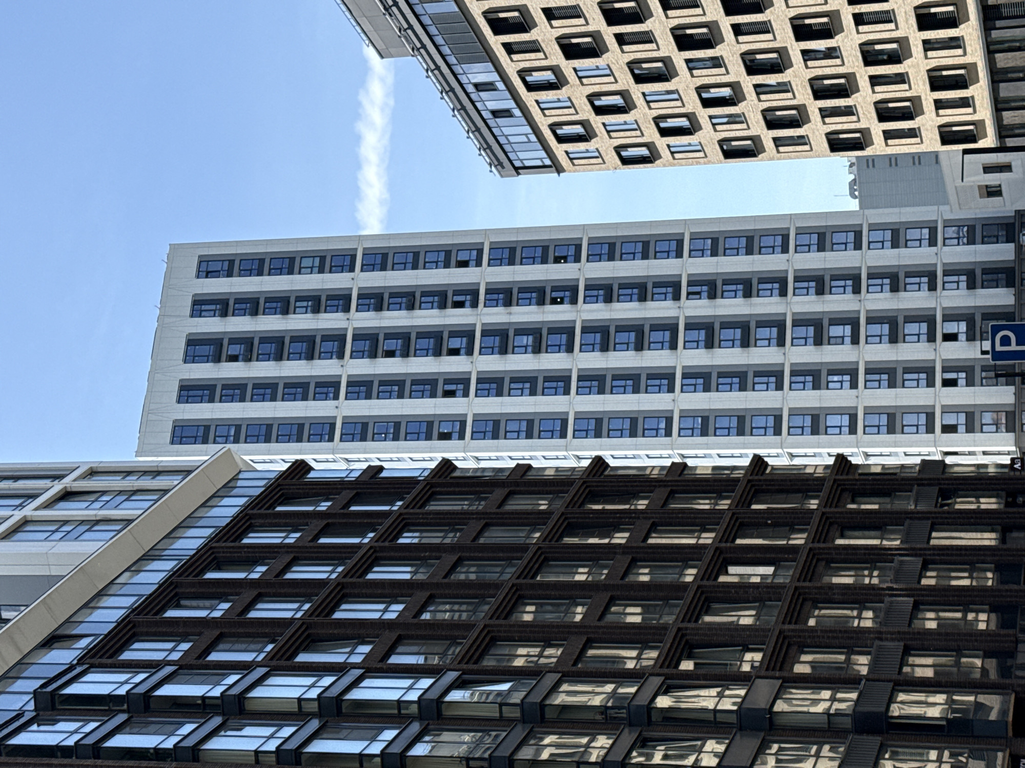 The image shows a low-angle view of three buildings in Moscow, Russia. The bottom building has a dark brown facade with square windows. The middle building is white with dark blue windows. The top building, partially visible, is tan with square windows. The sky is blue with a contrail. A parking sign is visible on the right.