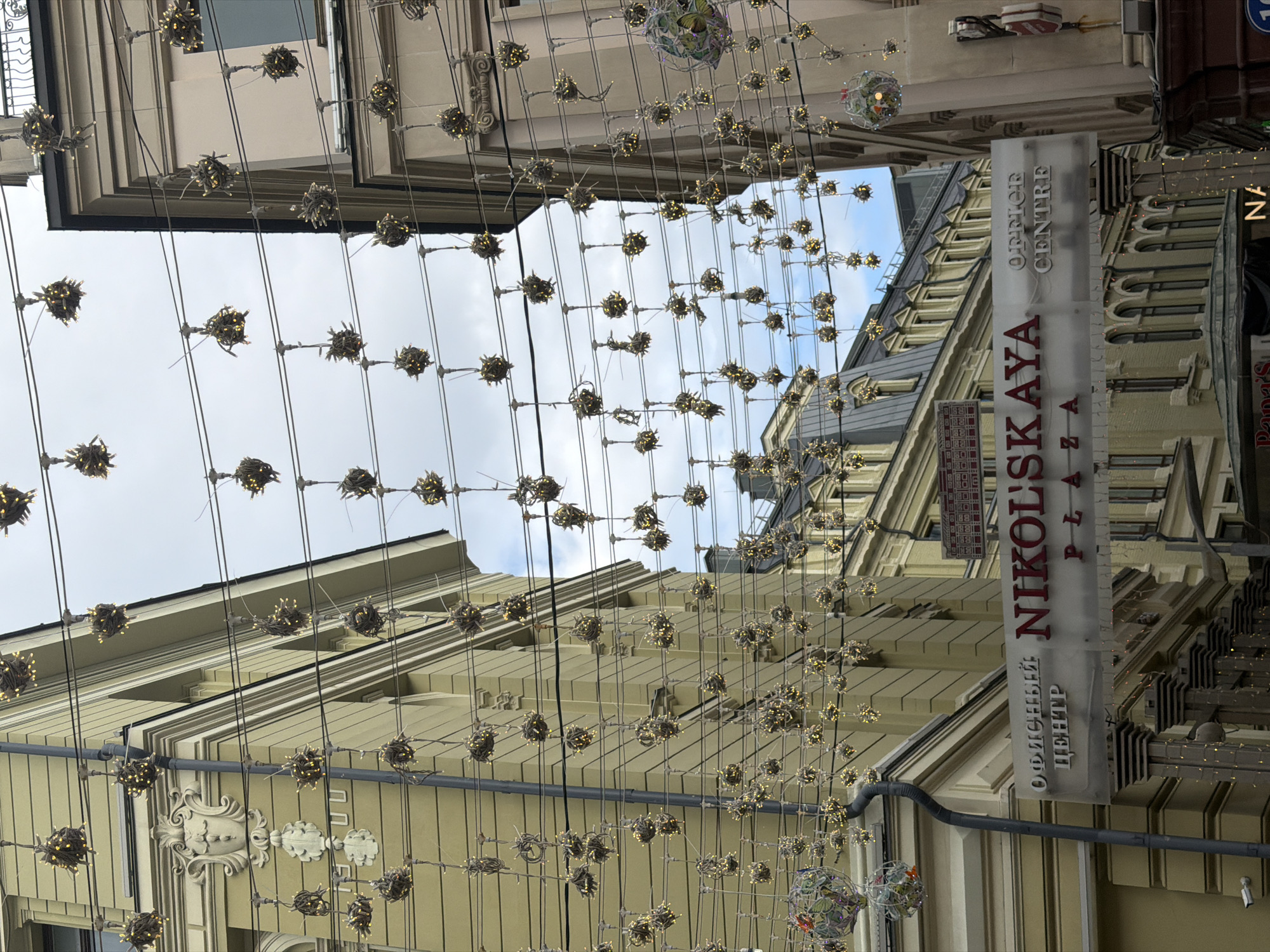 This image showcases a street in Moscow, Russia, characterized by ornate, light-colored buildings on either side, topped with decorative lighting suspended across the street. A sign for "Nikolskaya Plaza Office Centre" is visible on one side of the street, adding to the urban scene. The image's perspective is looking up, emphasizing the verticality of the buildings and the hanging lights. The sky is partially visible through the lighting, suggesting a bright, overcast day. There are no people visible in the frame.