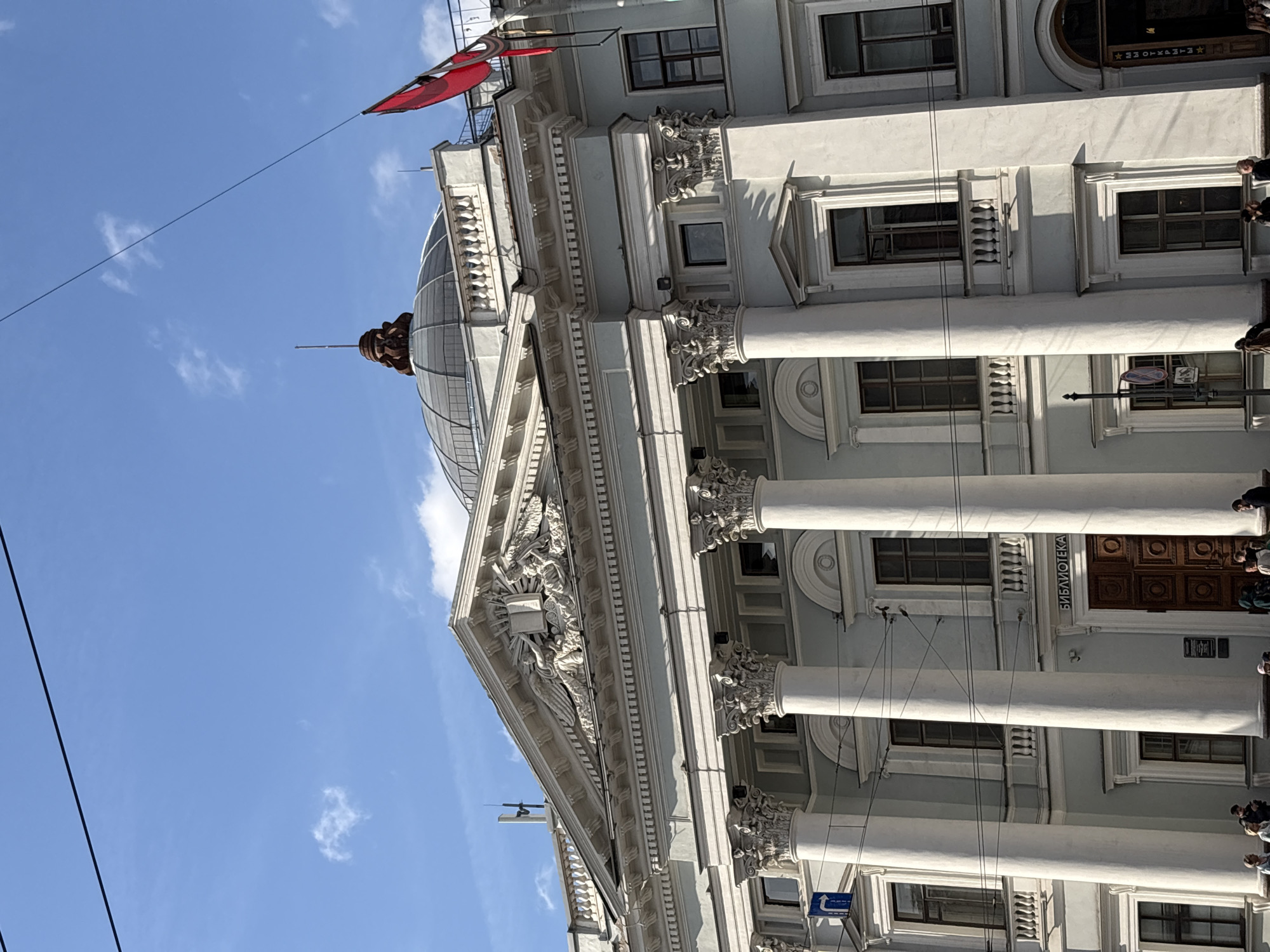 The image is an upward-angled view of a classical building with a dome and columns, set against a blue sky with scattered clouds. The building features ornate architectural details, including decorative carvings and a series of columns with Corinthian capitals. A red flag is visible at the top of the building. A sign with Cyrillic text on the facade reads "БИБЛИОТЕКА" (Library). Several people are standing near the building's entrance. The building is located in Saint Petersburg, Russia, specifically the Russian National Library.