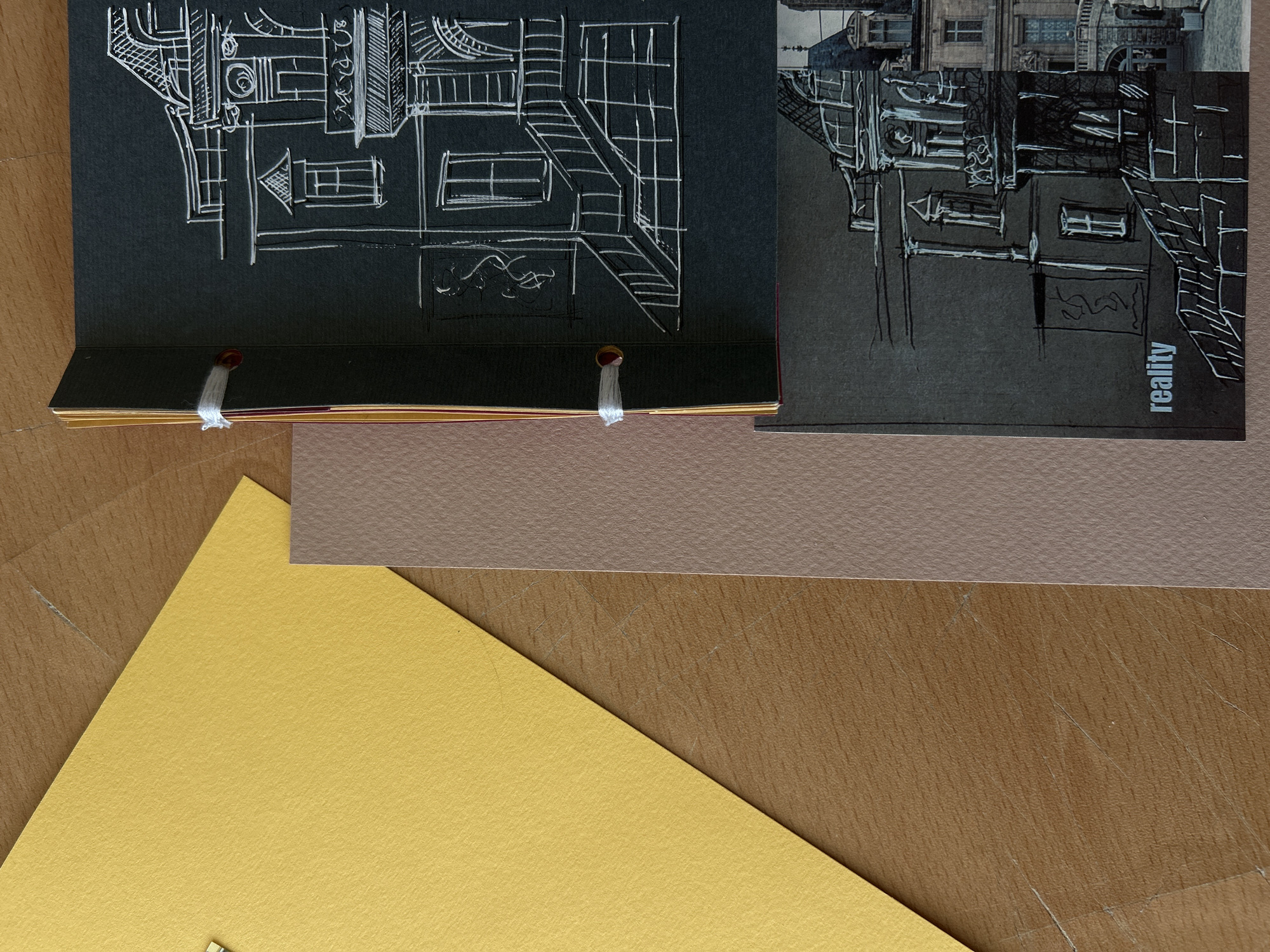 The image shows a close-up, top-down view of several pieces of paper arranged on a wooden surface. A bright yellow sheet of paper is positioned in the upper left quadrant, angled diagonally. To its right and slightly below is a narrow, vertical strip of light brown paper. Overlapping these elements and extending vertically down the center-right of the frame are two black sheets of paper, bound together with white string. The top black sheet features a white line drawing of a building facade with a staircase. The bottom black sheet has a similar line drawing of another building, with the word "reality" printed in white text in the lower left corner. The wooden surface appears to be a table with visible grain and some faint scratches. No people or actions are depicted, and no specific location in Sofia, Bulgaria can be identified.