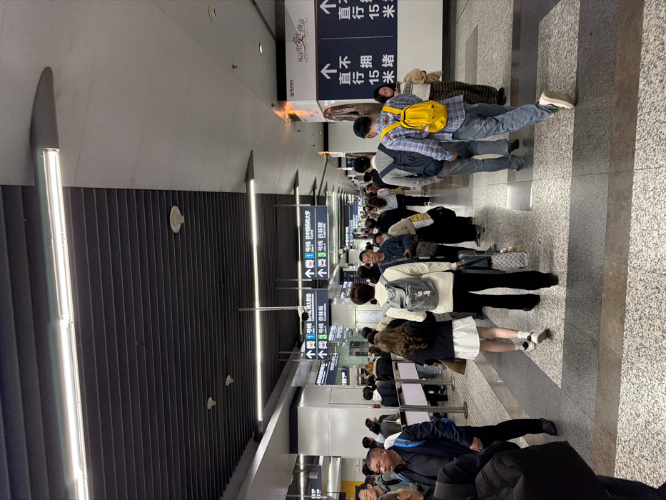 The image depicts a moderately busy underground transit station in Yuhuatai District, China. The environment is characterized by modern, functional architecture. The ceiling features dark gray slatted panels with embedded fluorescent strip lighting, interspersed with smoke detectors and fire suppression heads. Portions of the ceiling and upper walls are light gray. The flooring is a speckled light gray with dark gray rectangular pathway indicators.
Approximately two to three dozen individuals, primarily adults, are visible, engaged in transit activities. Most are walking in the direction away from the camera, though some are seen in profile or facing the camera in the mid-ground. Attire is predominantly casual, including sweaters, jackets, shirts, and jeans. Many individuals are carrying personal items such as backpacks and rolling luggage. A person in the foreground wears a white sweater and carries a light grey backpack and a patterned rolling suitcase. Another individual further back wears a plaid shirt and a bright yellow backpack.
Overhead, multiple white-on-blue directional signs with white arrows indicate routes for "1号线 Line 1" (Line 1) and "3号线 Line 3" (Line 3), specifying destinations such as "往 中国药科大学" (towards China Pharmaceutical University) and "往 林场" (towards Lincun). Along the right wall, a large informational or advertising panel displays an artistic graphic and Chinese text, including "直行 15 米" (go straight 15 meters). People are observed moving purposefully through the station, suggesting travel to or from platforms or exits. Interactions among individuals appear limited to walking in proximity.