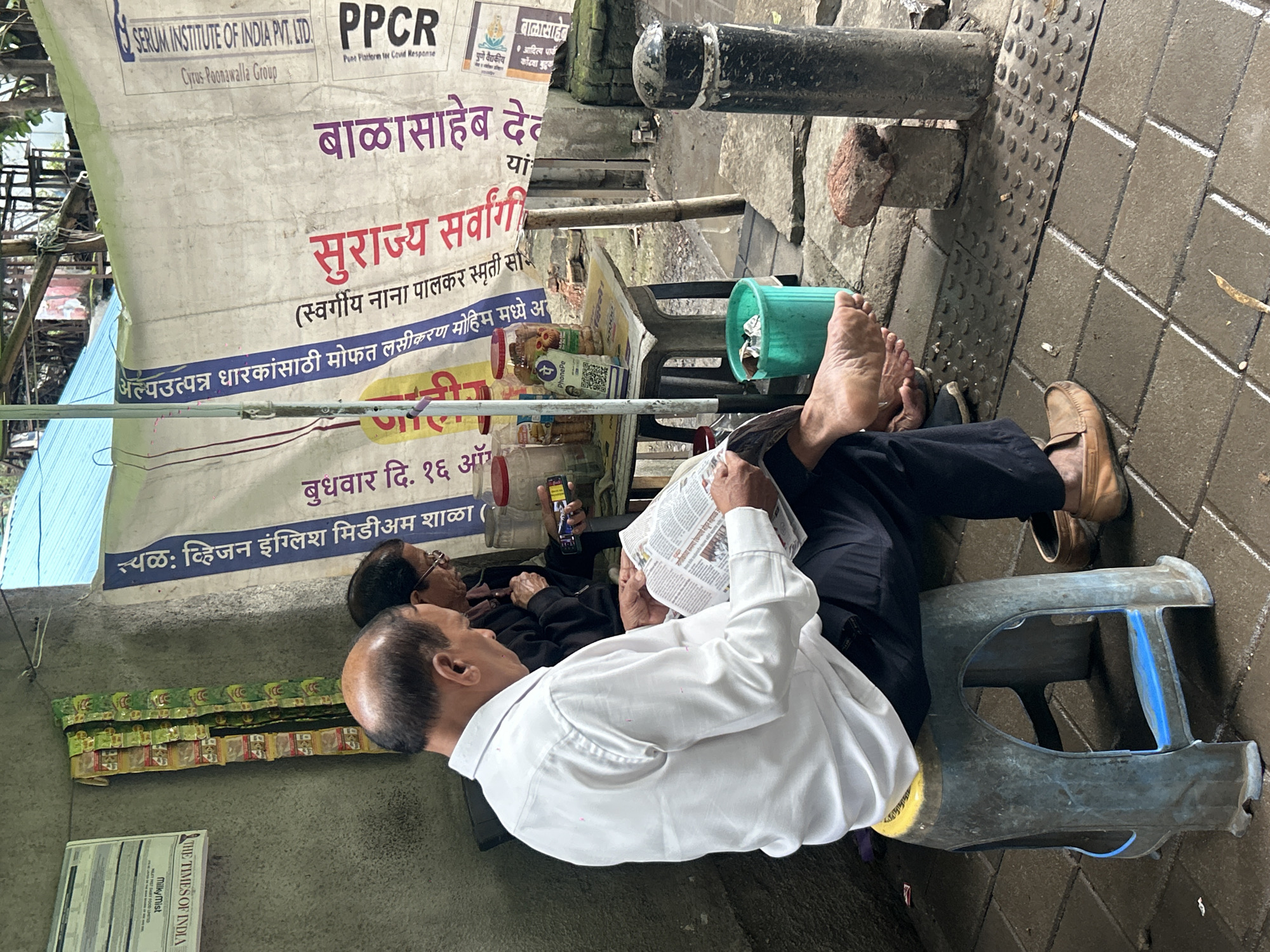 Two individuals, appearing to be adults, are seated outdoors in what resembles a marketplace or street-side vendor area. The person in the foreground, a male with a bald head, is wearing a white shirt and dark trousers. He is seated on a blue plastic stool and is engrossed in reading a newspaper. His bare feet are visible, resting on the ground. Beside him, to the right, another individual, possibly female, with glasses, is also seated and appears to be holding a mobile phone.
The environment suggests a casual, possibly informal commercial setting. Behind the individuals, a table displays various items including jars filled with what might be snacks or other consumables, and a green trash bin. A large banner with text in what appears to be Marathi script hangs behind the table. The ground is paved with textured tiles, and the overall lighting indicates it is daytime. The presence of the banner with text, the variety of goods on display, and the paved public space suggest an urban setting in Pune, India. The banner's text is partially visible, and some English text is also present, including "SERUM INSTITUTE OF INDIA PVT. LTD." and "PPCR". The poster to the left appears to be a newspaper, "THE TIMES OF INDIA". The overall scene depicts a moment of pause and activity in a busy urban environment.