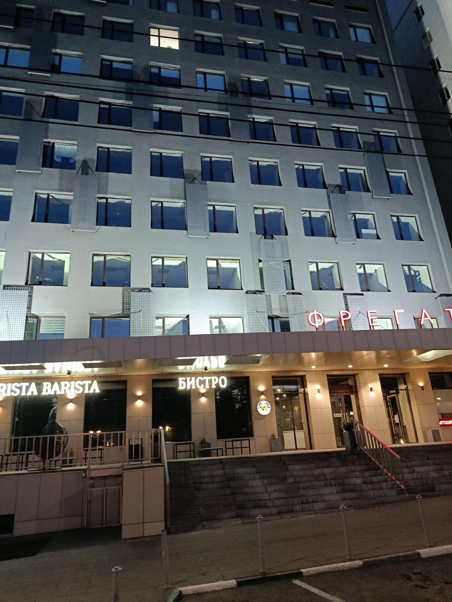 A multi-story building is illuminated at night, with a sign reading "FRIST BARISTA" and another reading "БИСТРО" (BISTRO) visible at the lower level. The building features a repeating pattern of windows and balconies. A staircase leads up to the entrance of the establishments, and a sidewalk with white markings is visible in the foreground. The overall scene appears to be a commercial area within the city of Saratov, Russia, based on the Russian text present. There are no people clearly visible in the image.