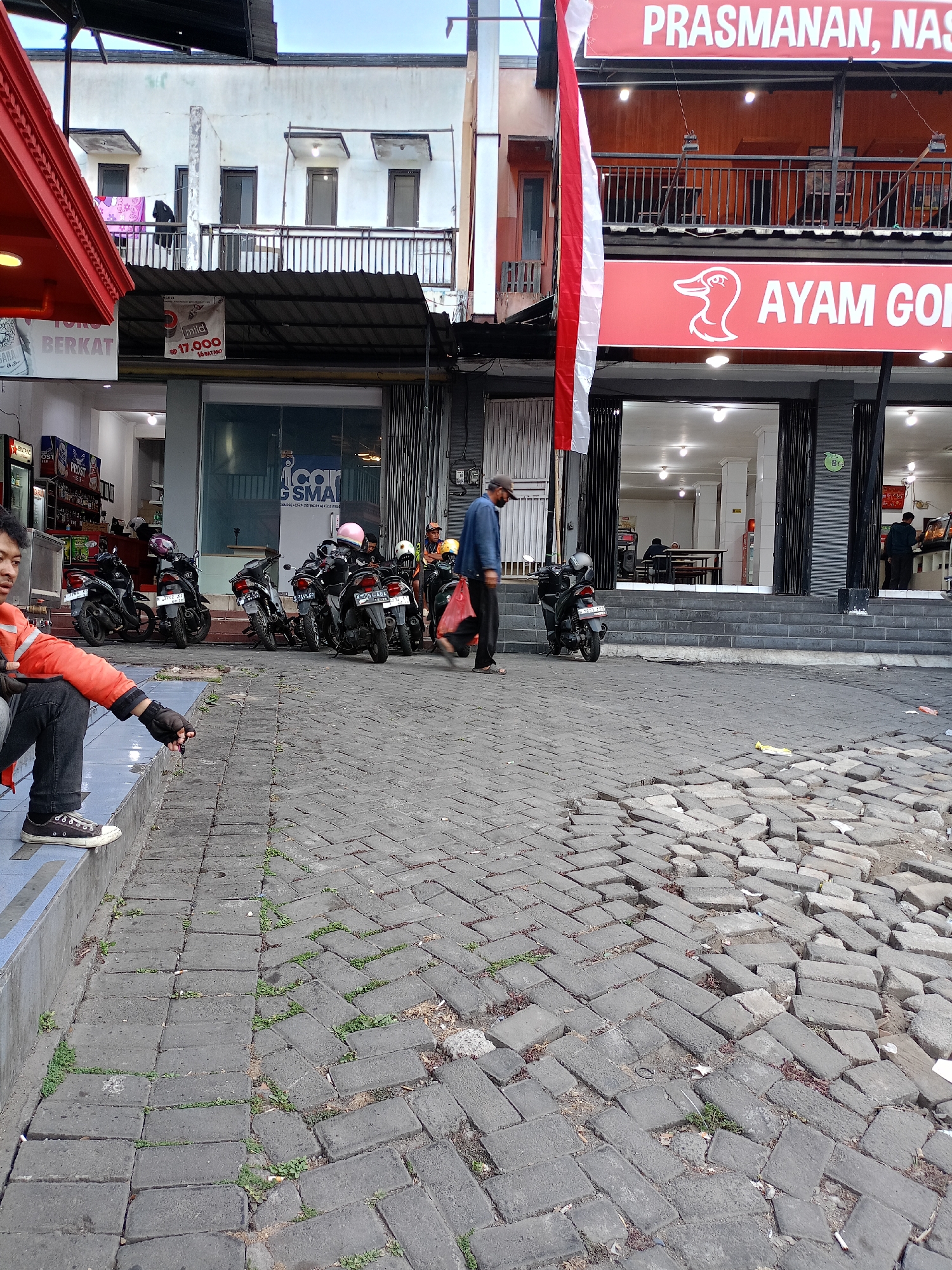 The image captures a street scene in Batu, Indonesia, featuring a row of parked motorcycles in front of a building. A person in an orange jacket is seated on a blue step on the left, and another person in a blue shirt and cap is walking away from the camera towards the right. The building appears to be a commercial establishment, possibly a restaurant, with signage including "AYAM GORENG" (Fried Chicken). The street is paved with cobblestones, some of which are displaced, indicating wear and tear. Greenery is visible growing in patches between the cobblestones. The overall atmosphere suggests a busy urban environment, possibly during daylight hours given the lighting.