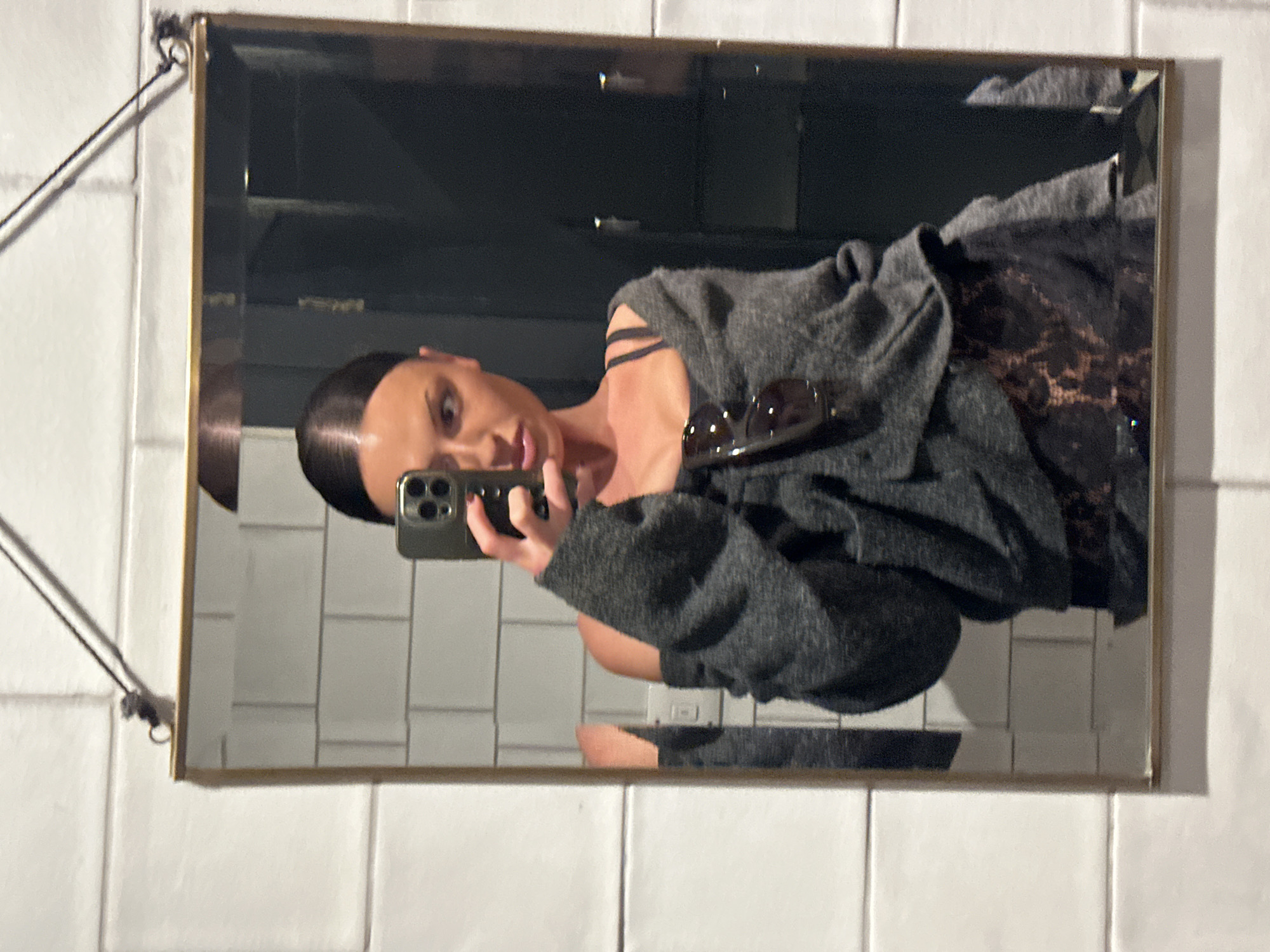 An image captures a mirror selfie of a woman taken indoors. The woman is reflected in a rectangular mirror with a brass-toned frame, which hangs by a dark cord against a white tiled wall. She has dark hair pulled back neatly and is holding a dark-colored smartphone with multiple camera lenses in her left hand to take the photo.
She is wearing a dark top, possibly patterned or lace, under a grey, textured cardigan or jacket. A pair of dark sunglasses is resting on her chest or the cardigan. Her gaze is directed towards the phone, indicating she is posing for the selfie. The background reflected in the mirror behind the woman is dark and indistinct. The white tiled wall, with its grid pattern, is clearly visible around the mirror and in reflections within it. The lighting suggests an artificial indoor environment. The image is oriented sideways, presenting the woman horizontally. No visible text is present. The location context provided is London, United Kingdom.