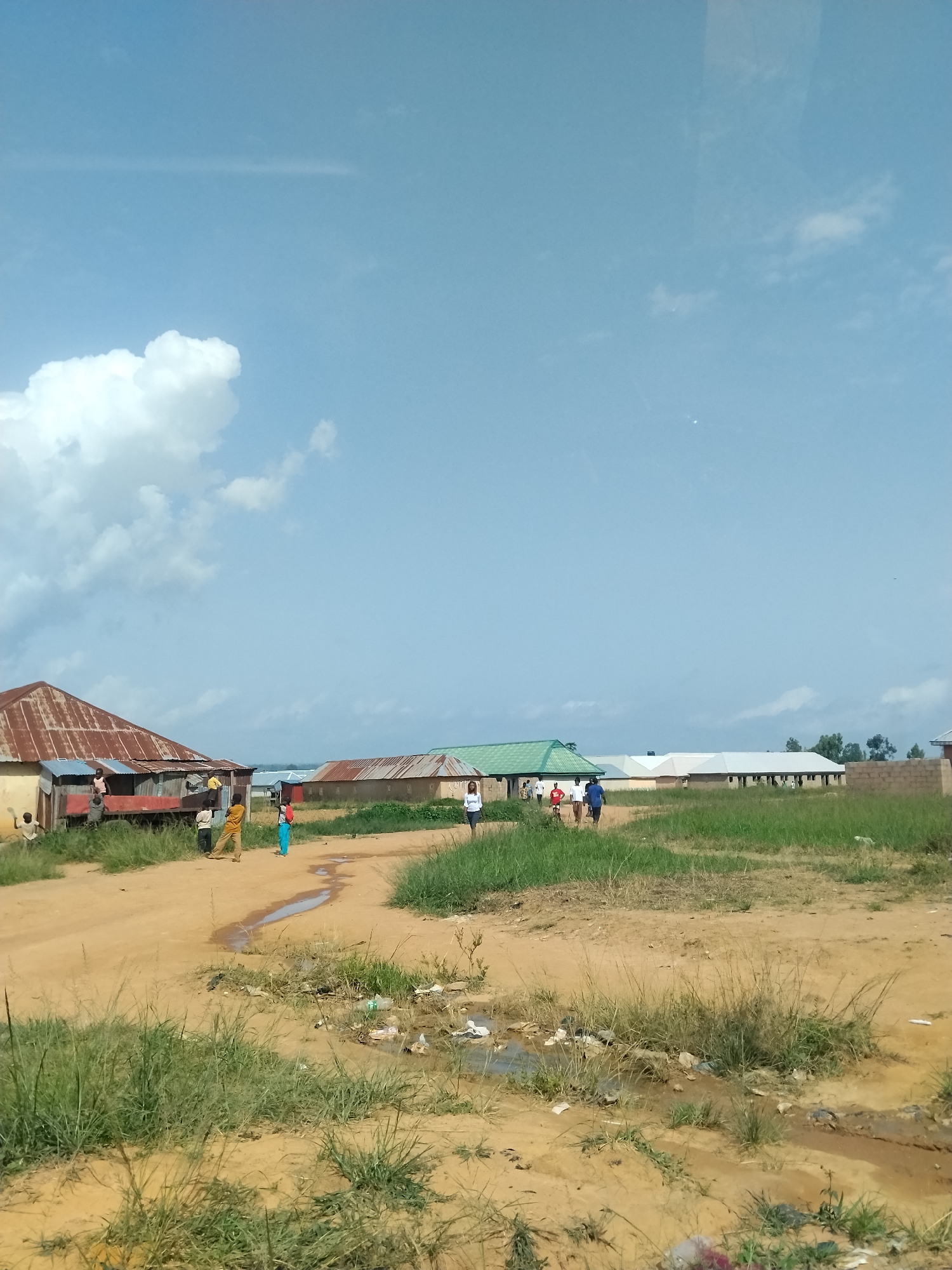 The image depicts a scene in Durumi, Nigeria, under a partly cloudy sky. In the foreground, a dirt road with puddles and scattered debris, including plastic bags, is visible, bordered by overgrown grass. Several people are present: a group of children are playing near a rusty-roofed building on the left, some standing and others interacting. To the right of the road, more individuals are walking, including a woman in a white top and dark skirt. Behind them, a row of buildings with corrugated metal roofs and some with green roofing material stretches into the distance. The environment appears to be a residential or semi-rural area with basic structures.