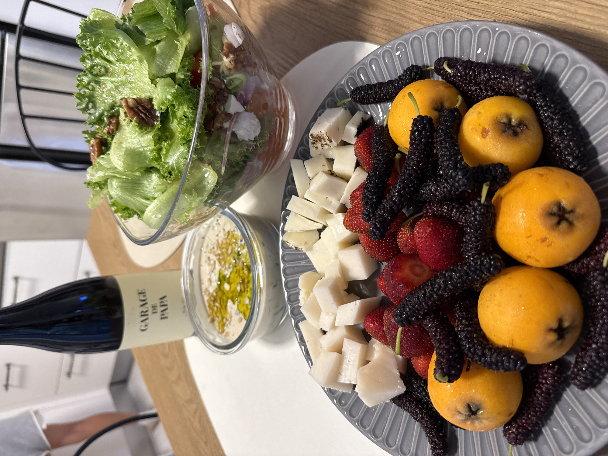 On a light-colored surface, likely a tabletop, there is an arrangement of food items and a bottle of wine, suggesting a meal setting. To the left, a bottle of wine labeled "GARAGE DE PAPA" is positioned near a glass bowl filled with a green salad, including lettuce and what appear to be nuts. Adjacent to the salad is a small bowl filled with a dip, possibly hummus, drizzled with olive oil.
To the right, a grey decorative plate contains an assortment of fresh items: cubed white cheese, red strawberries, dark purple mulberries, and several bright yellow loquat fruits. The arrangement is dense and colorful, suggestive of a selection of appetizers or a dessert platter.
The setting appears to be indoors, possibly in a home dining area, given the casual arrangement and the presence of kitchen appliances visible in the background. The image was taken in the city of Herzliya, Israel.