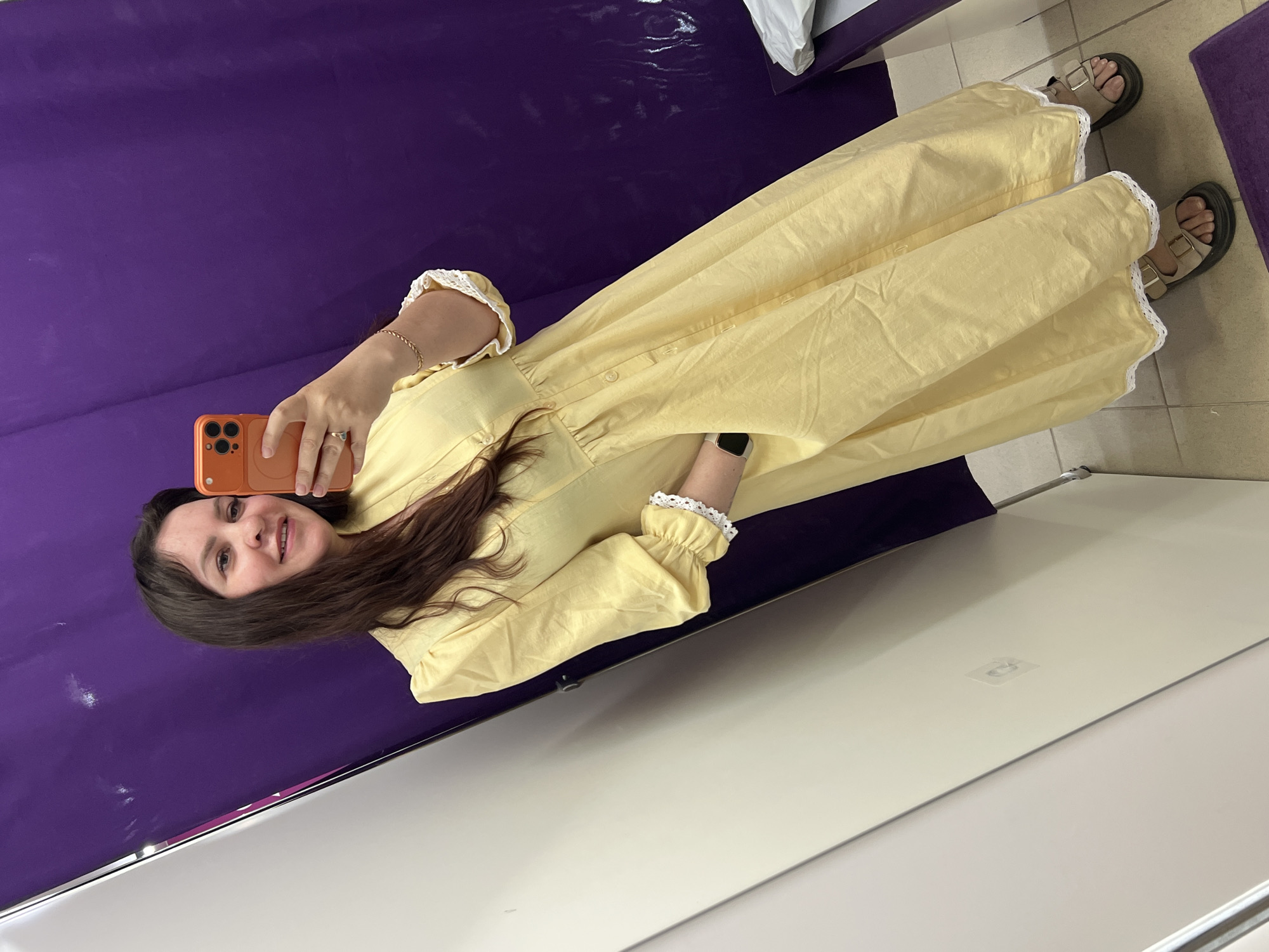 A woman in a yellow dress with lace trim on the sleeves and hemline is taking a selfie using a phone with an orange case. She is standing in front of a purple backdrop. She is wearing sandals. The location is in Noviy, Russia.