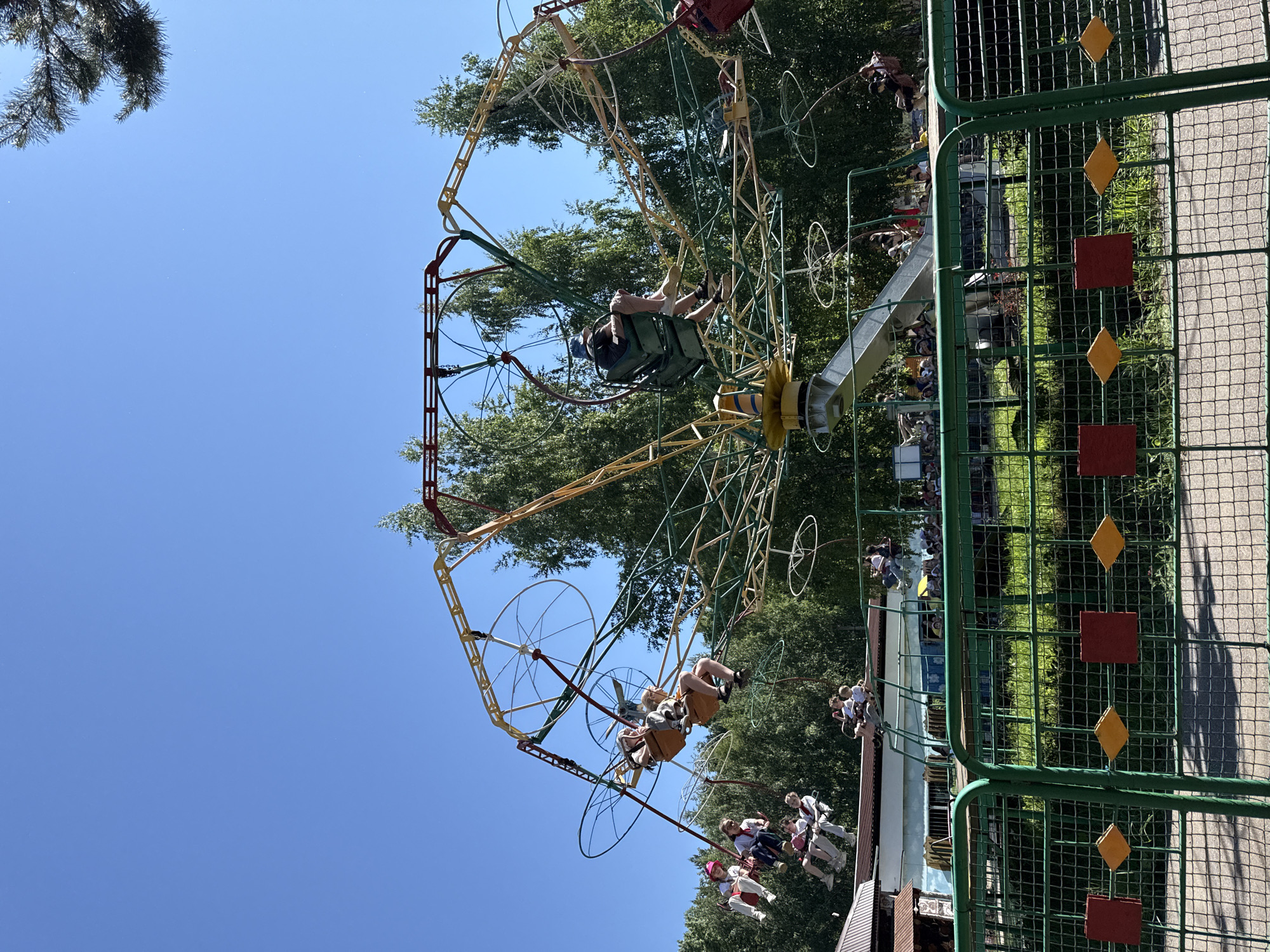 The image depicts an amusement park ride in Chaikovsky, Russia, under a clear blue sky. The ride, a circular swing-like attraction, is in motion, with several people seated in suspended carriages. The ride's structure is made of metal, painted in yellow, green, and red. Trees surround the attraction, and a green metal fence with red and yellow diamond-shaped decorations runs across the foreground. The ride appears to be operational, with children and adults enjoying the experience. The overall scene suggests a sunny day at a recreational facility.