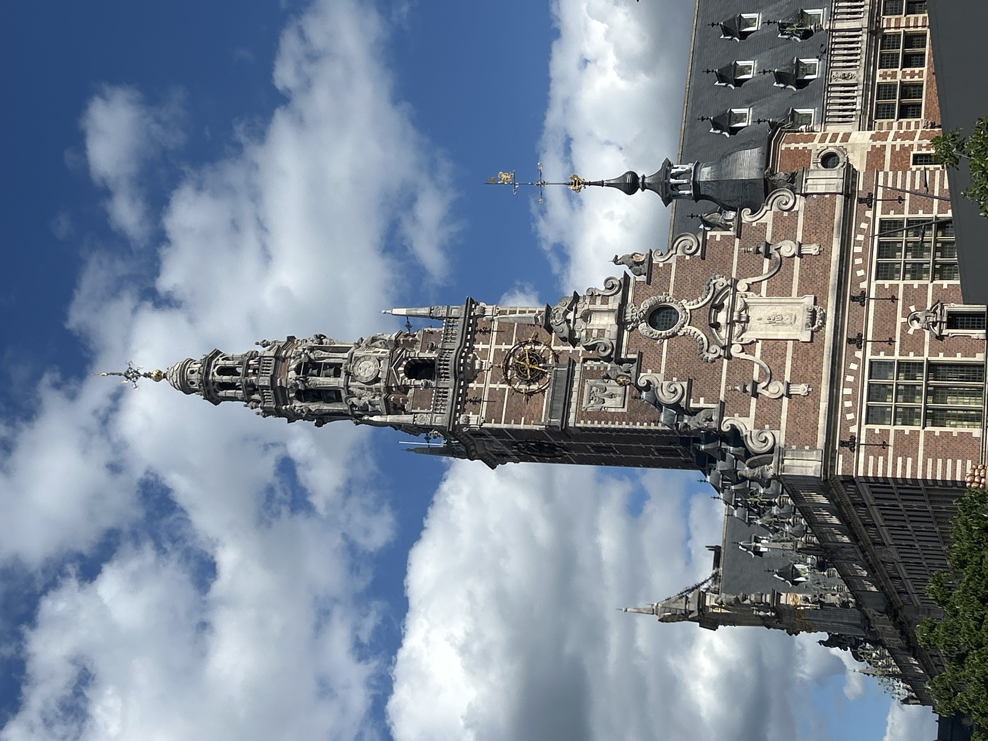 The image shows a close-up of the University Library of Leuven in Belgium, a historic building with a prominent clock tower. The tower is made of brick and stone, with intricate architectural details and a tall spire topped with a weather vane. The building features multiple windows, decorative carvings, and a large clock face on the tower. The sky is partly cloudy, with a bright blue background and white cumulus clouds. In the foreground, the tops of trees are visible. The overall scene is a detailed view of the architectural grandeur of the University Library.