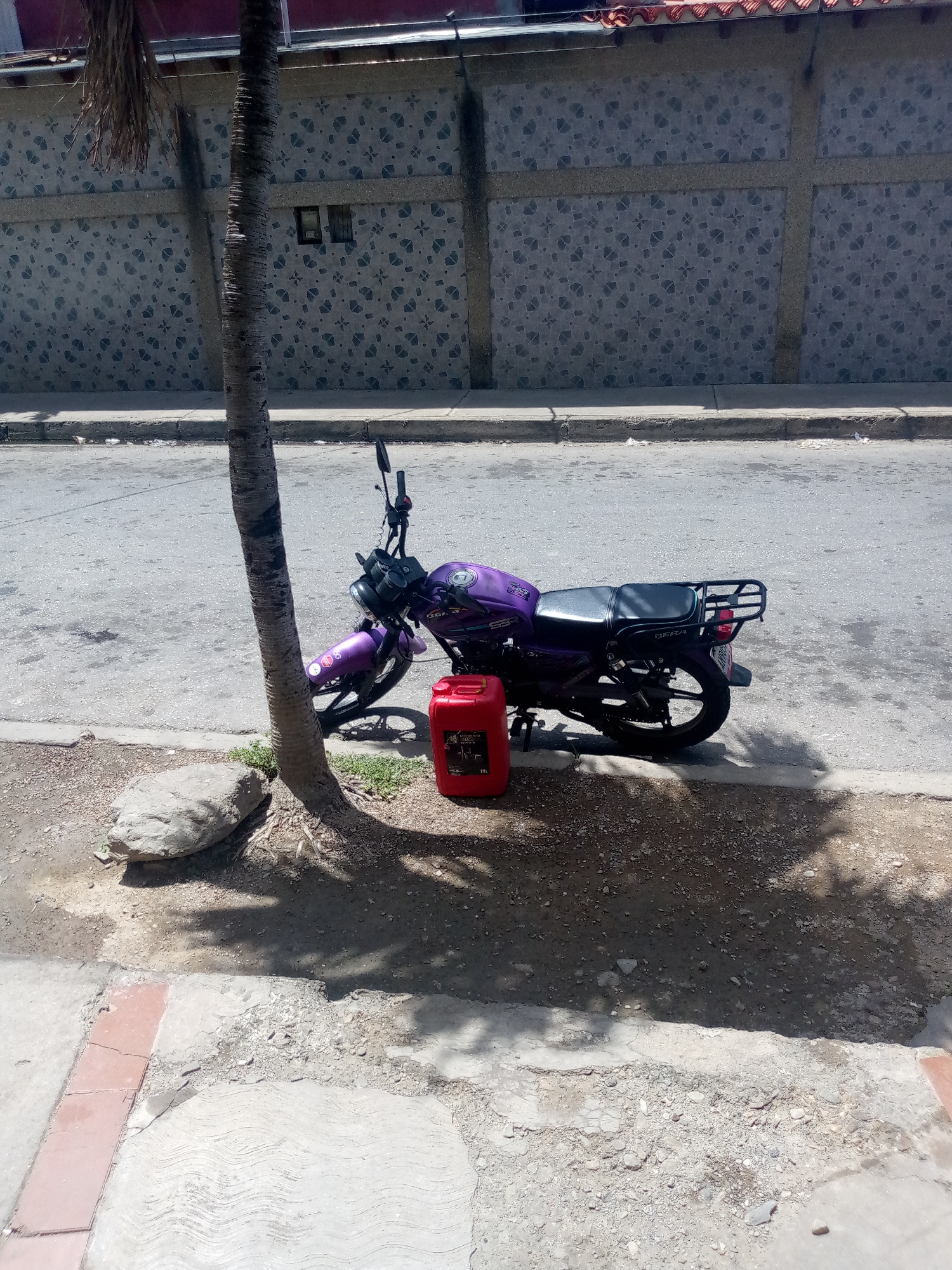 A purple motorcycle is parked on the side of a street, next to a palm tree. A red plastic gas can is placed on the ground in front of the motorcycle. The motorcycle has a black seat and a black rack on the back. The gas can appears to have some text on it, but it is not legible. The scene is outdoors, and the lighting suggests it is daytime, possibly mid-morning or afternoon, with shadows cast by the tree and what appears to be a building. The ground is a mix of asphalt and dirt, with some rocks and vegetation visible. A concrete sidewalk runs along the side of the street. In the background, there is a wall with a patterned design. The overall scene is a typical street view in a sunny, possibly warm climate. There are no people visible in the image.
