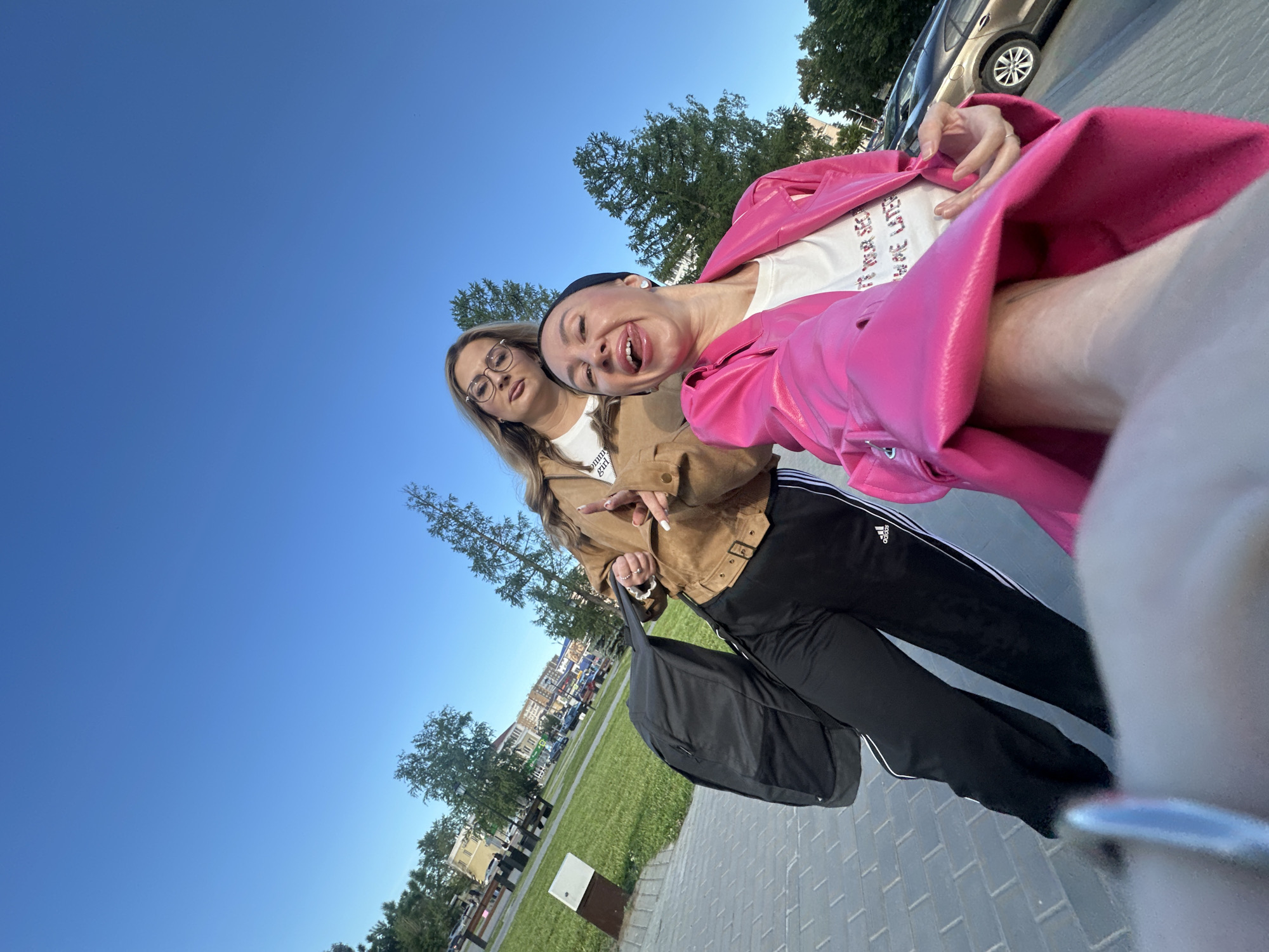 Two young women are depicted in a low-angle, tilted outdoor photograph, likely a selfie, under a clear, bright blue sky in Izhevsk, Russia. The strong sunlight suggests it is daytime, possibly late afternoon.
On the right side of the frame, one woman with dark hair pulled back by a black headband playfully sticks out her tongue and has a wide, open-mouthed smile. She is wearing a vibrant, glossy pink jacket over a white t-shirt. Visible text on her t-shirt reads, "I'LL SEE YOUR SEX AND RAISE YOU LATER." She also wears black track pants with distinctive white Adidas stripes and a logo on the outer seam of the left leg.
To her left, the second woman, wearing glasses and a light brown jacket, poses with a more subtle expression, looking slightly off-camera. A white t-shirt is visible beneath her jacket, with partially visible text that appears to say "TOMMY GIRL." She has long, light brown hair styled in braids or plaits. Her left hand holds a black shoulder bag, while her right hand is raised, making a "peace" sign (two fingers up) and showcasing light-colored nail polish.
The background features a paved sidewalk in the foreground, transitioning to a grassy area with several trees. In the distance, buildings and parked cars are visible, indicating an urban or park-like setting. The overall mood is casual and playful.