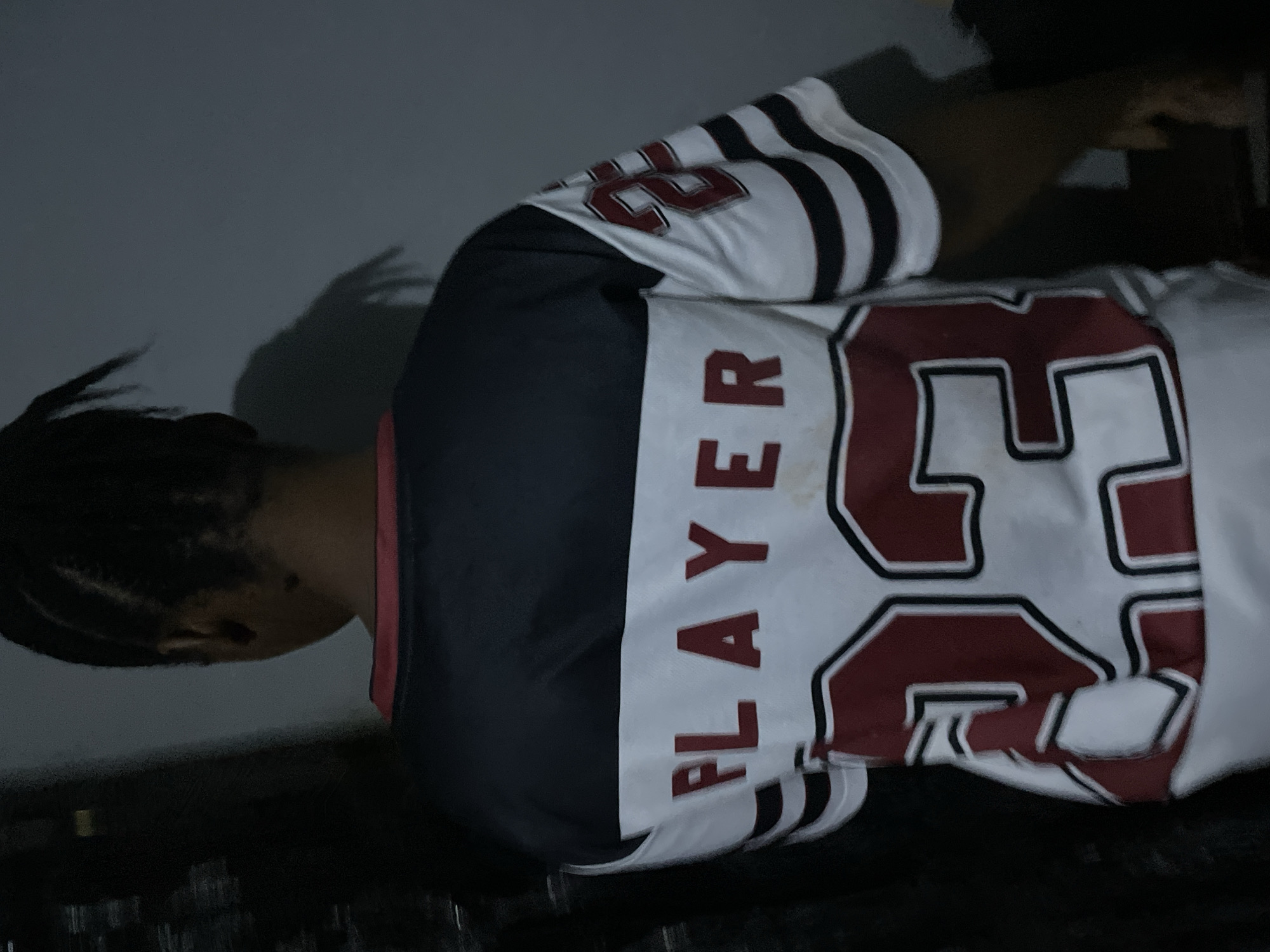 A person is wearing a white and black athletic jersey with the word "PLAYER" vertically aligned on the left side and the number "23" prominently displayed on the front and back. The jersey has striped sleeves. The person's hair is braided. The background includes a light-colored wall and an indistinct dark area, possibly a piece of furniture. The image lacks specific details to determine if it was taken in Jos, Nigeria.