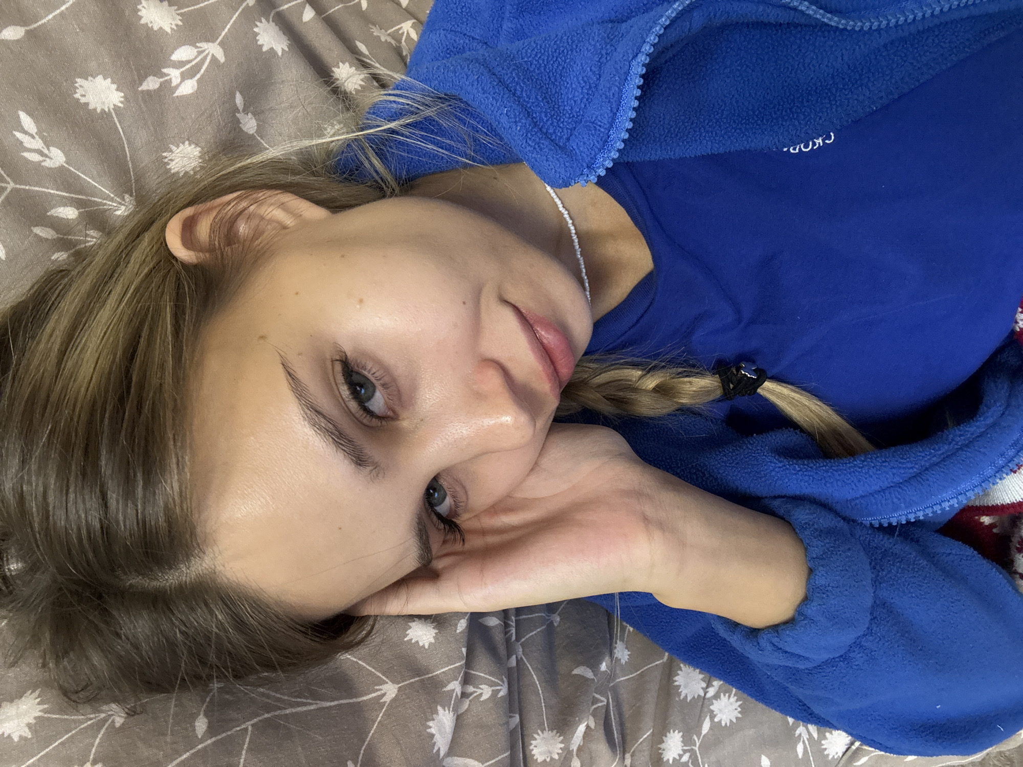 A fair-skinned young woman with light brown hair lies on a patterned bedspread with a floral motif. She is wearing a blue fleece jacket over a blue shirt. Her hair is styled in a braid, secured with a black elastic. Her head rests on her right hand. The word "CKOD" is partially visible on her shirt.
The location is not discernible from the image. Thus, no location in the city of Chaikovsky, Russia can be ascertained.
