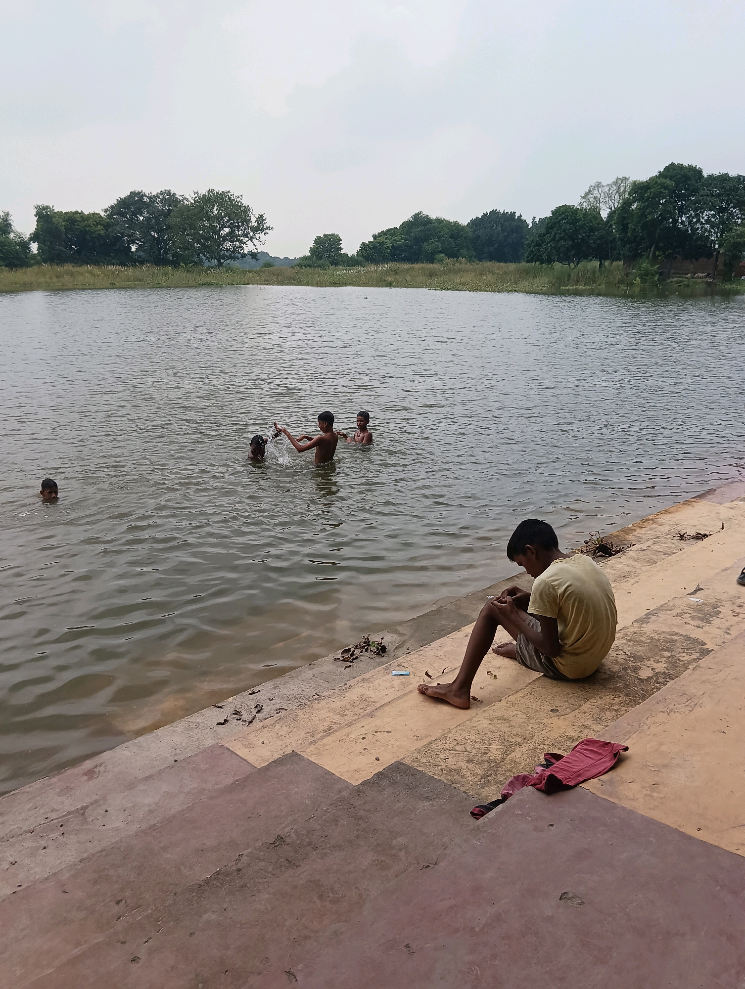 In Tilkar, India, a serene body of water serves as a backdrop for a group of boys. Four boys are in the water, with one of them splashing. To the right of the water, on a concrete embankment, a boy sits alone, looking down at something in his hands. His shirt is a light yellow, and he is wearing gray shorts. A red cloth is laid out on the embankment near him. The environment is natural, with trees visible in the background. The sky is overcast.