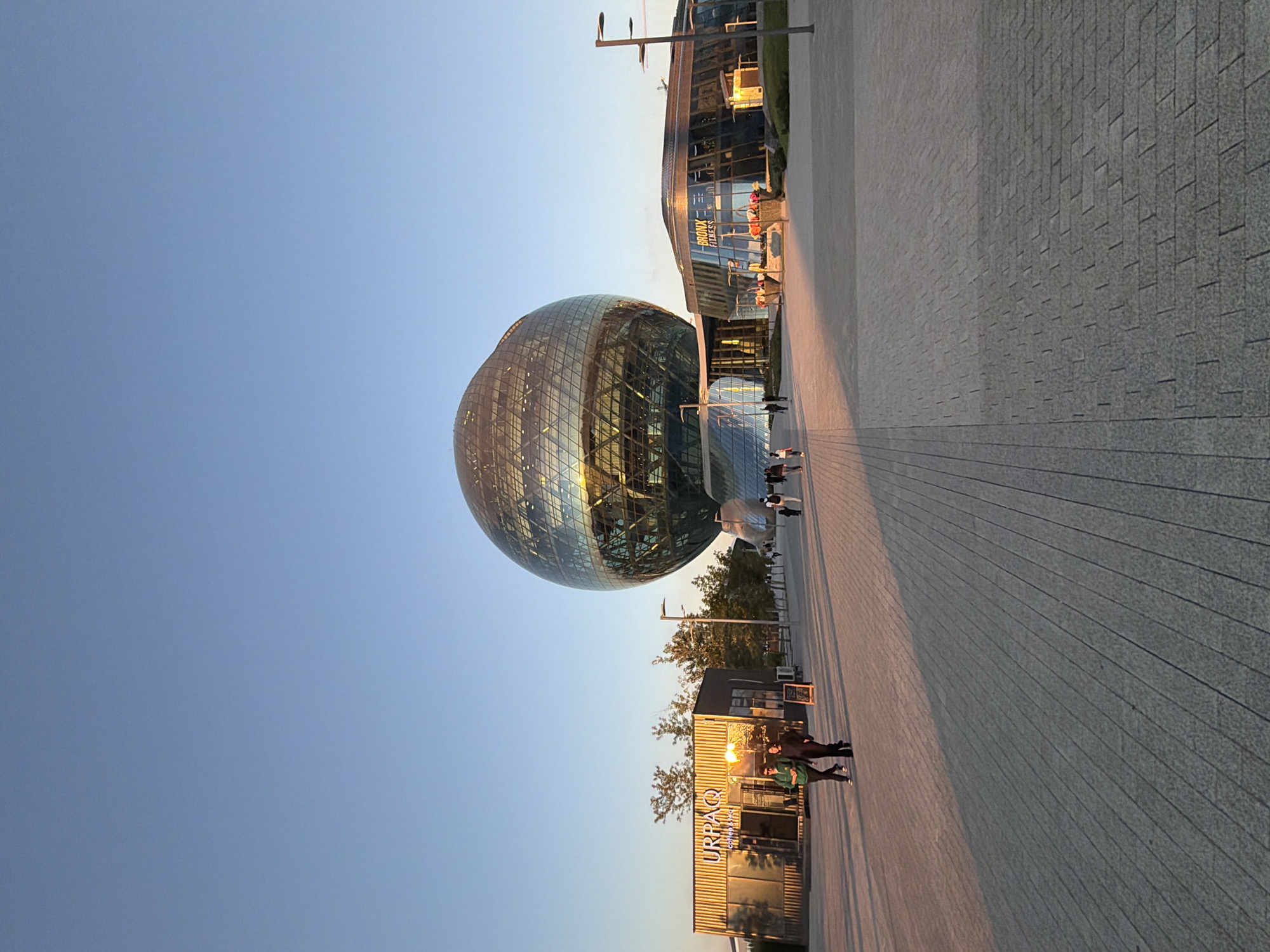 The image captures a wide-angle view of the Khan Shatyr Entertainment Center in Astana, Kazakhstan, under a clear, pale blue sky. The iconic spherical building, clad in a diamond-patterned glass facade, dominates the center of the frame, reflecting the warm, golden light of what appears to be late afternoon or early evening. To the left of the main structure, a smaller, modern building with signage reading "URPAQ COFFEE&FOOD" is visible, with two individuals walking in front of it. Further to the right, another contemporary building with glass walls and signage for "BRONX FITNESS" is partially visible. The foreground consists of a wide, paved walkway made of interlocking gray pavers, with long shadows cast across it, indicating the low angle of the sun. Several lampposts are present along the walkway. In the background, trees with sparse foliage line the area, and other people are seen walking at a distance. The overall scene conveys an open, public space with modern architectural elements bathed in soft, natural light.